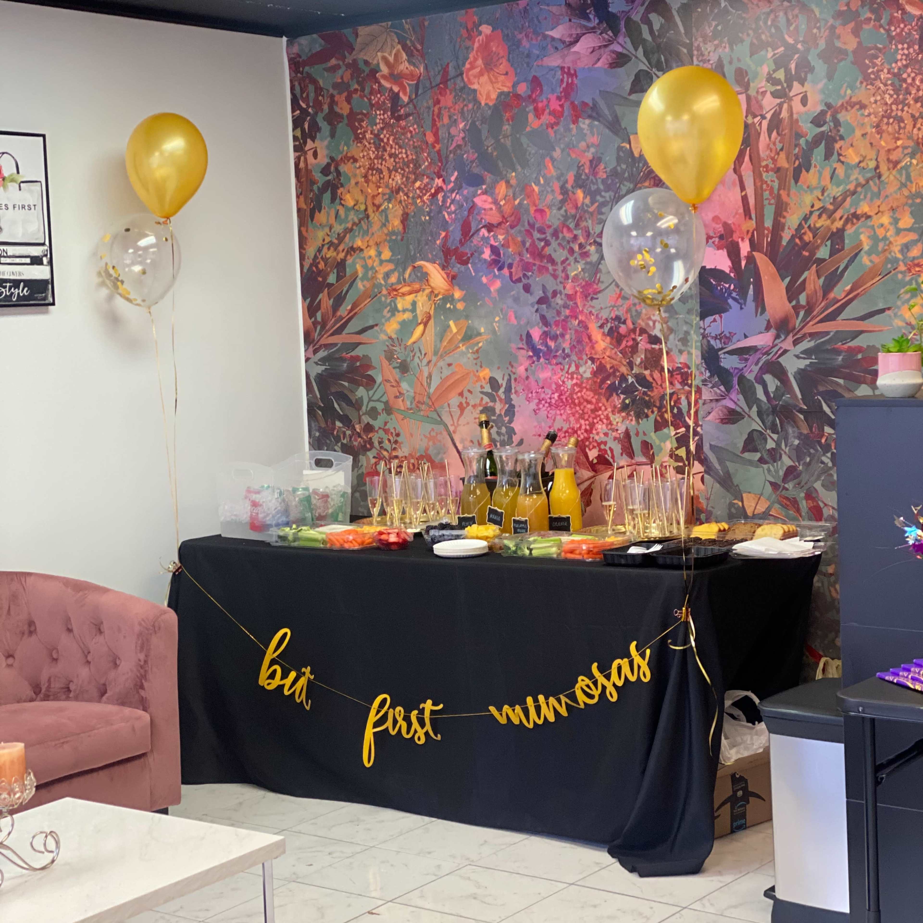 A party setup features a decorated table with snacks, drinks, and balloons against a vibrant floral backdrop.