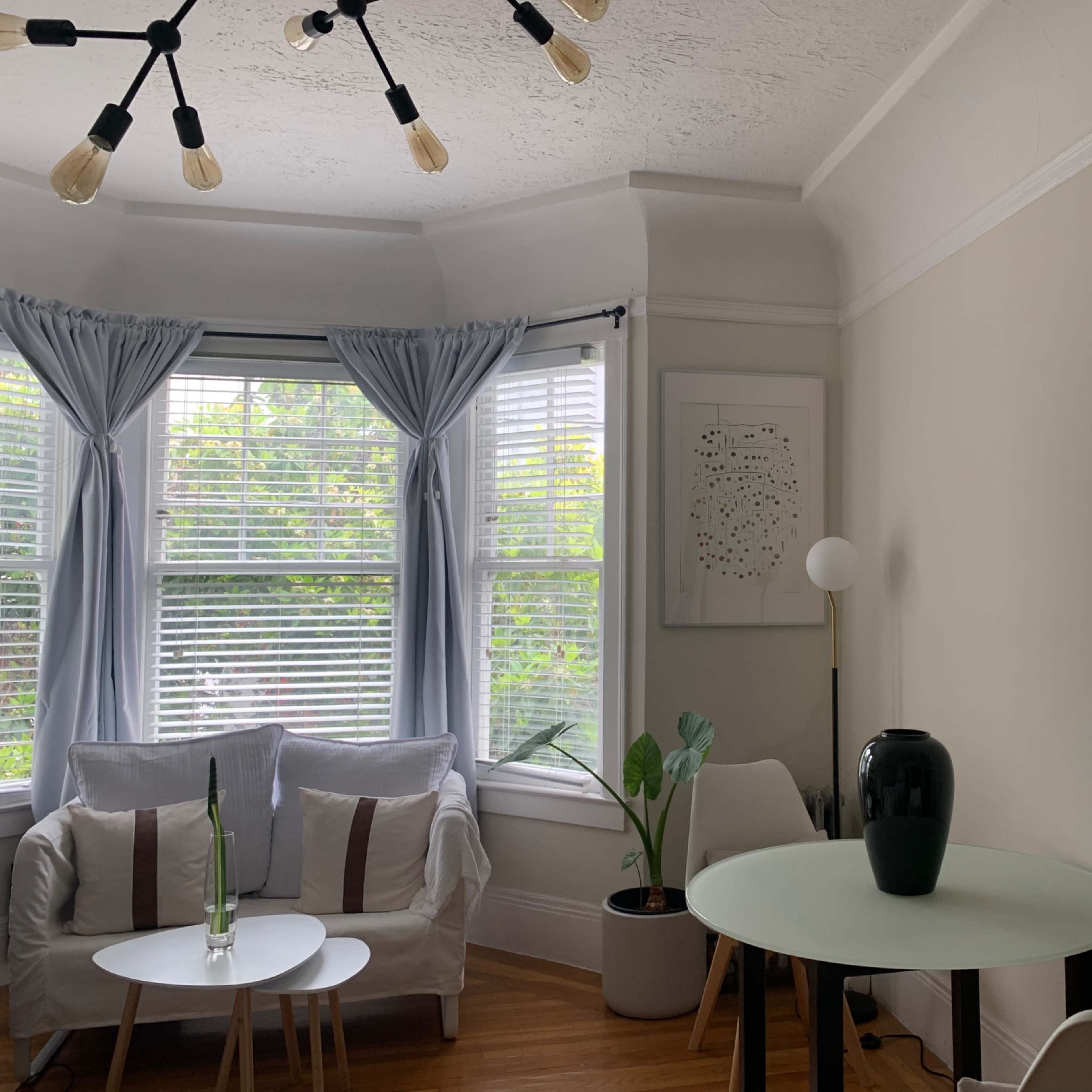 A living room with large windows, a white couch, a round table, a floor lamp, a potted plant, and a wall mirror.