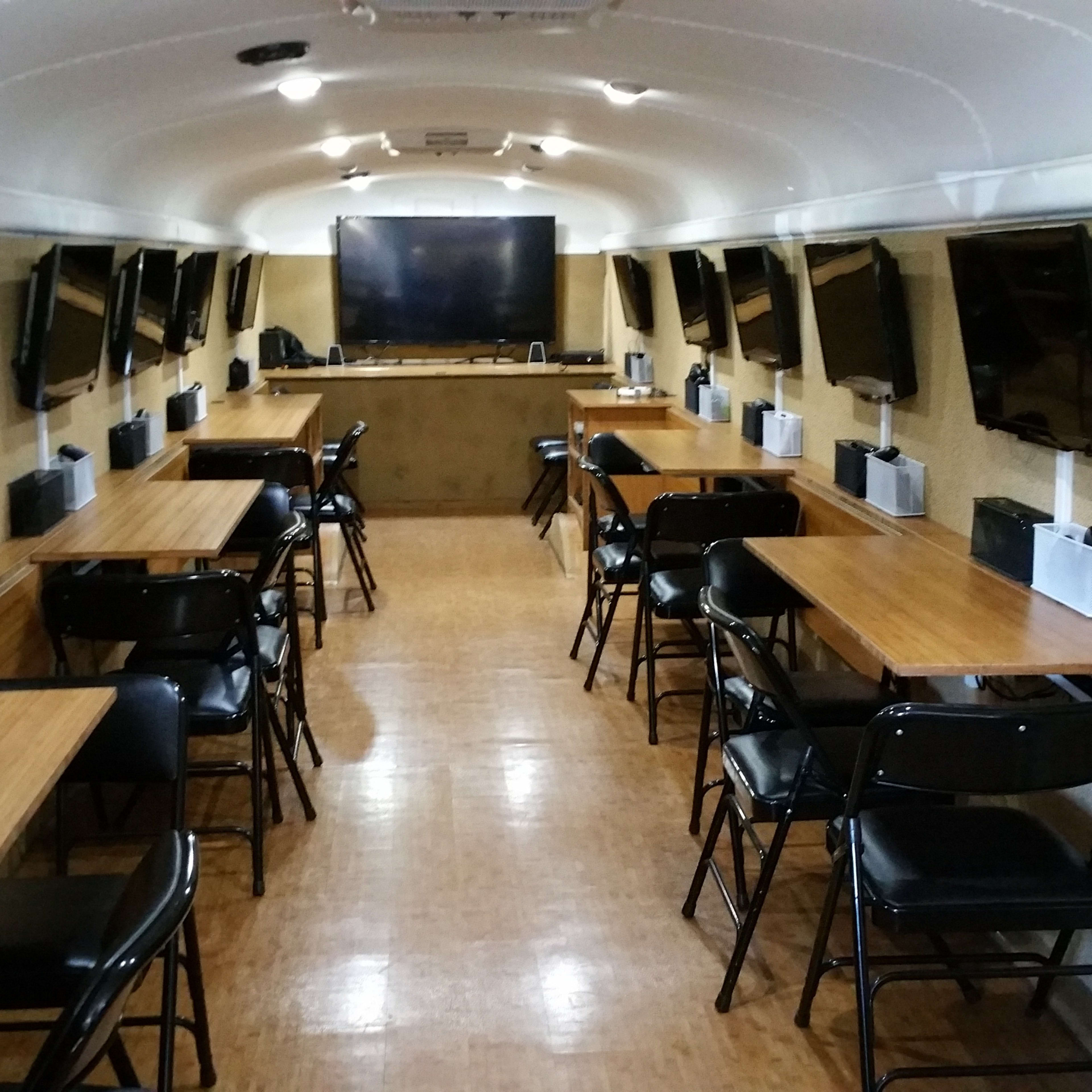 The interior of a bus is equipped with multiple flat-screen TVs, rows of wooden tables, and black chairs arranged for a group setting.