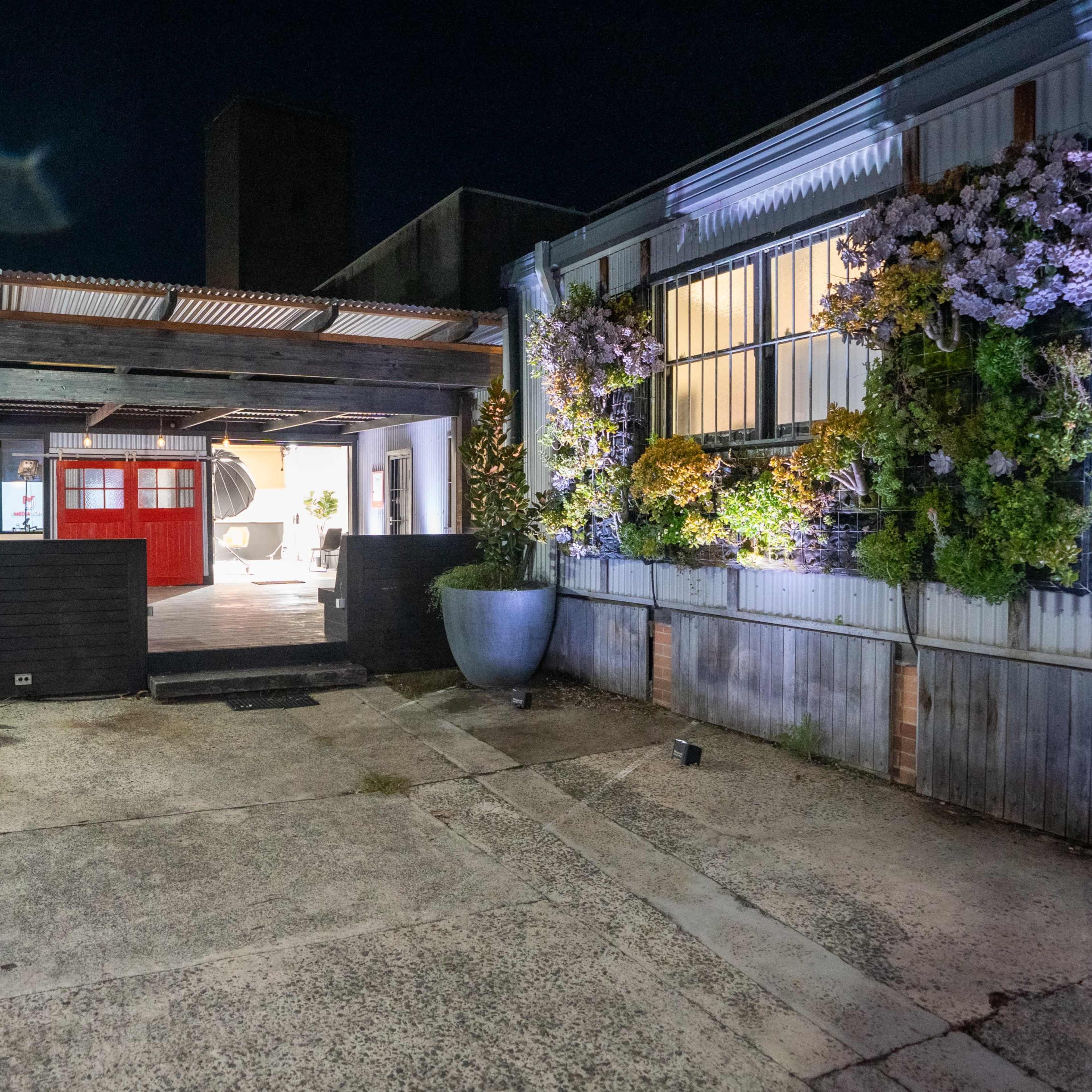 A narrow courtyard at night features a mix of metal walls and greenery, with illuminated windows revealing a red door in the background.