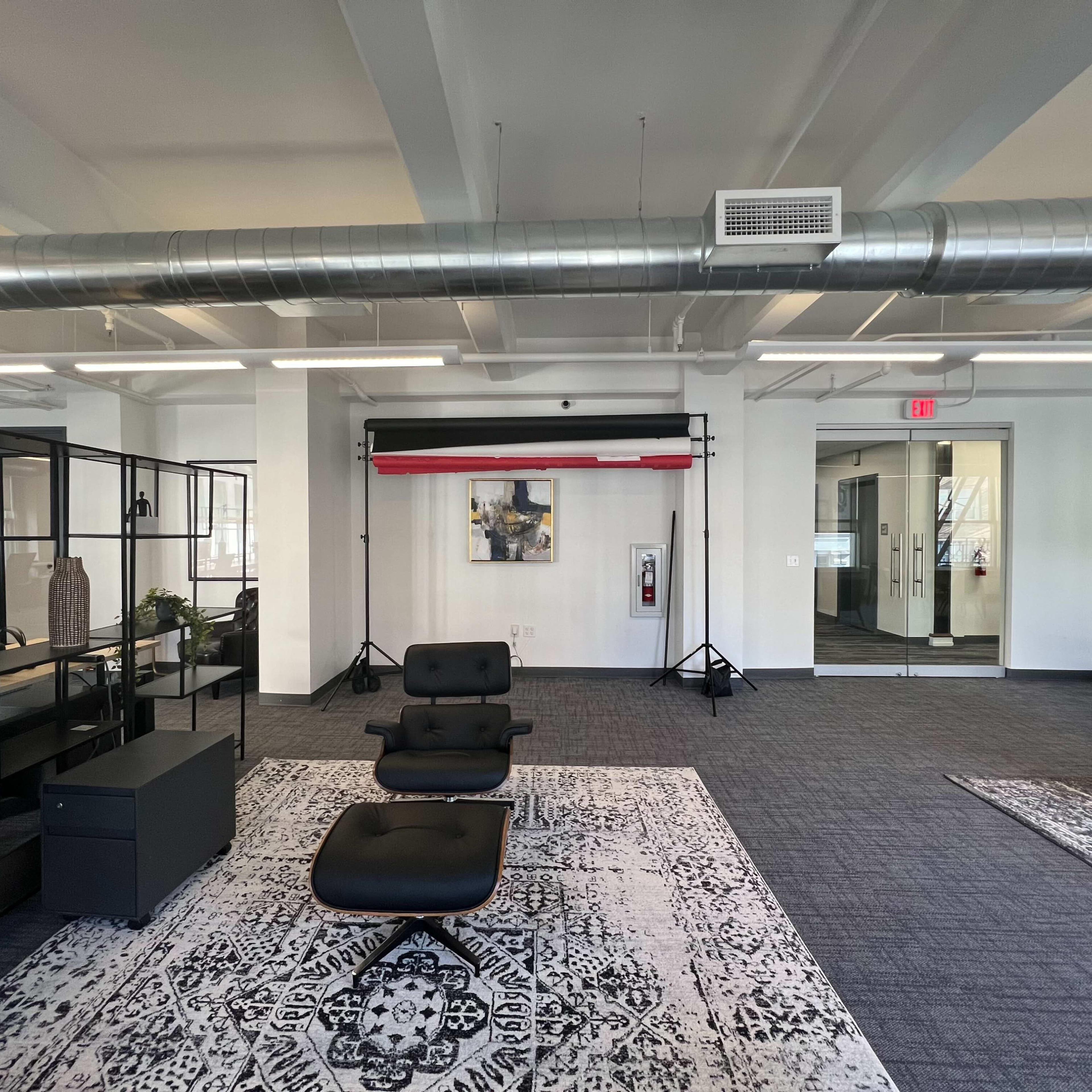 A modern office space with black furniture, a patterned rug, and a backdrop setup near a glass-walled meeting room.