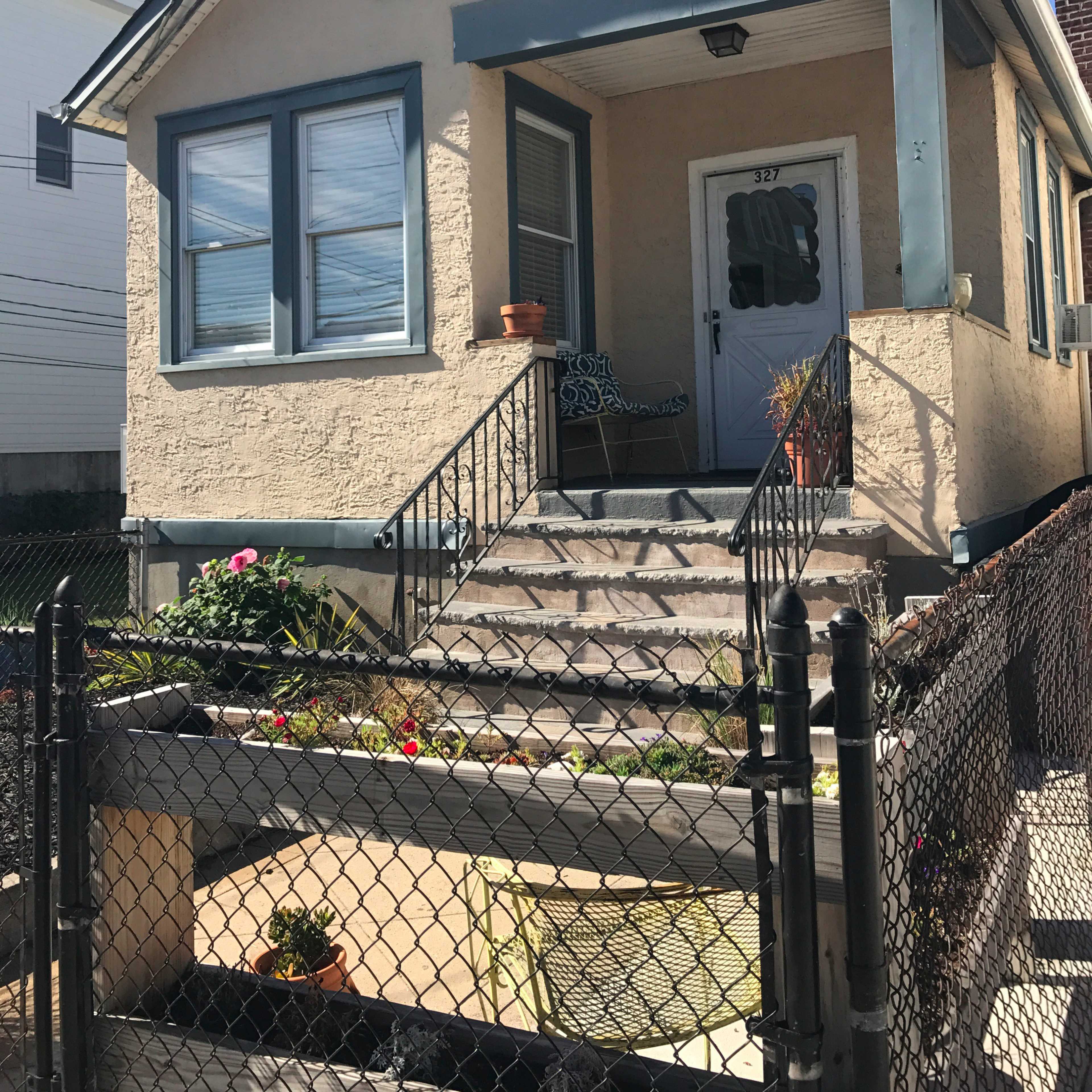 A small, single-story house with a front porch, surrounded by a fence and featuring a garden area.