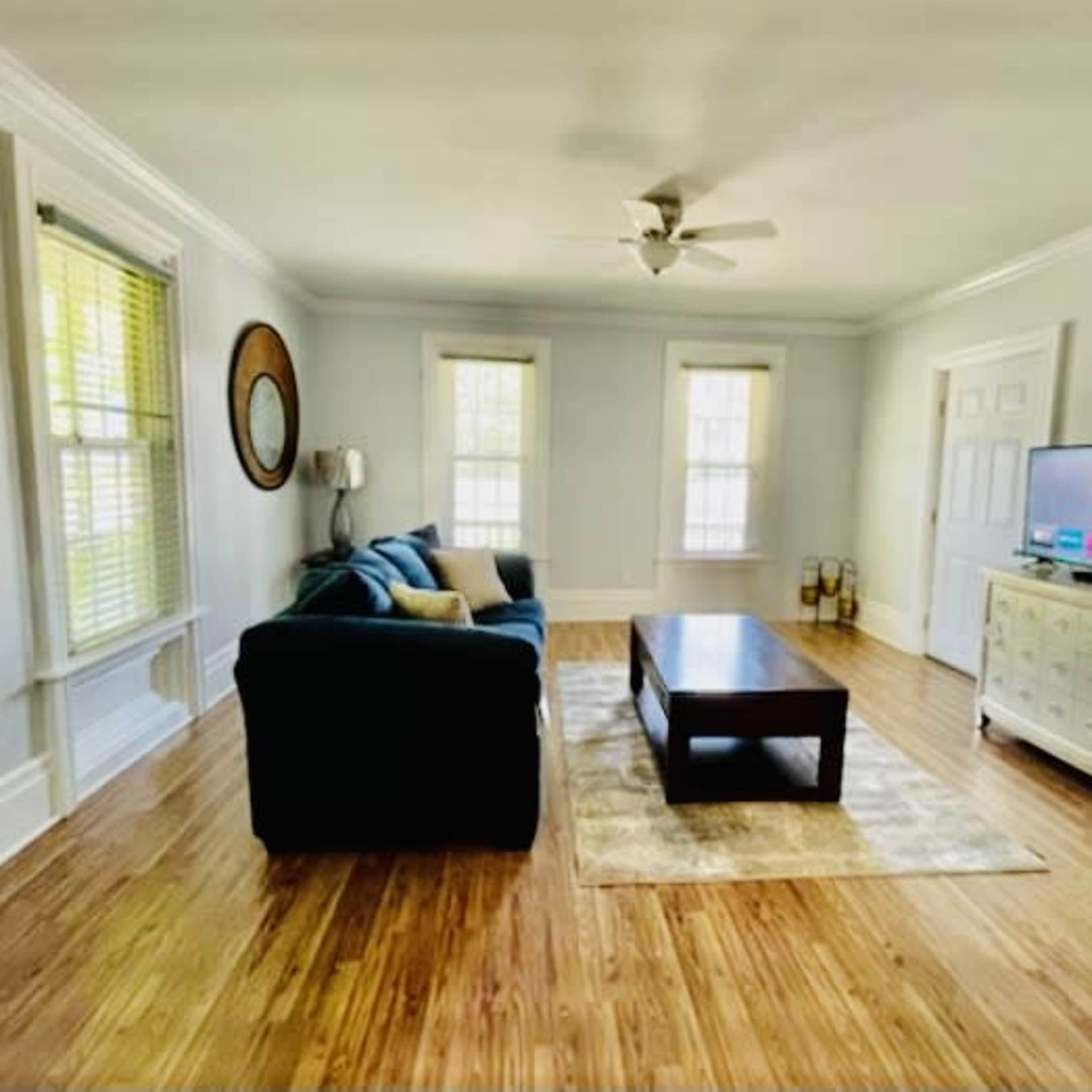 The room features a blue sofa, a wooden coffee table, and a television, with large windows allowing natural light to illuminate the space.