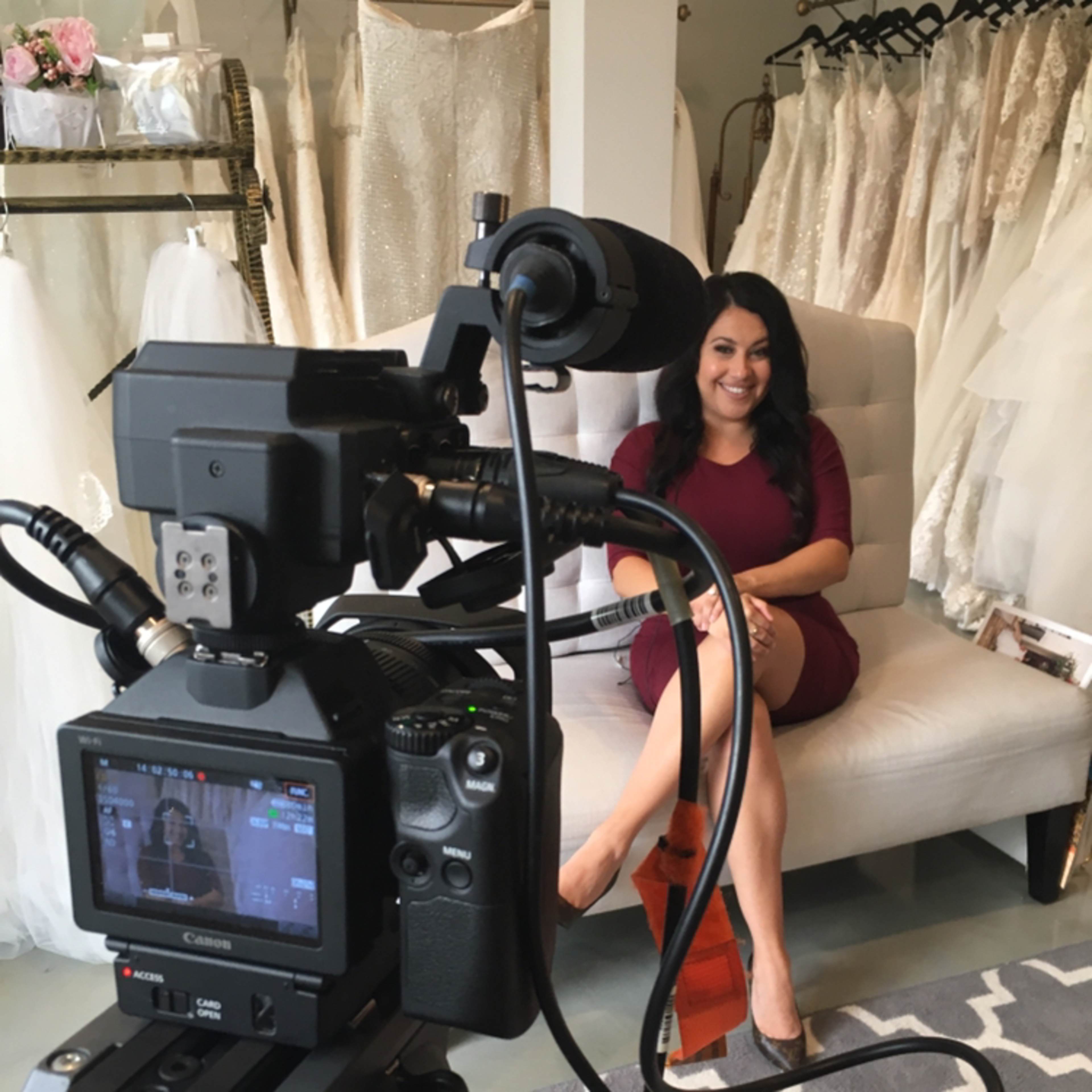 A camera is set up in a bridal shop, focusing on a woman sitting on a couch among wedding dresses on display.