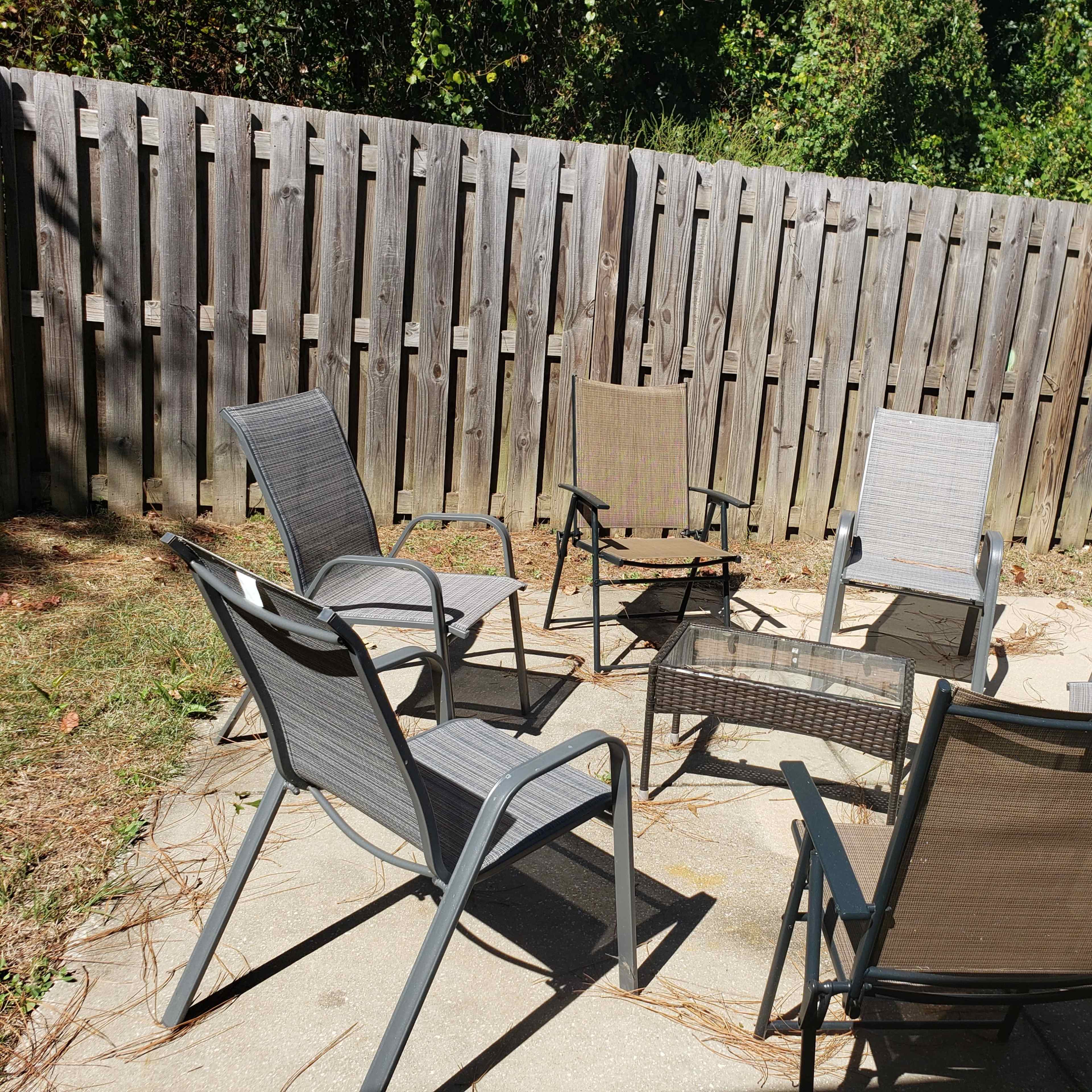 A circular arrangement of six patio chairs and a central glass table is situated on a concrete patio surrounded by a wooden fence.