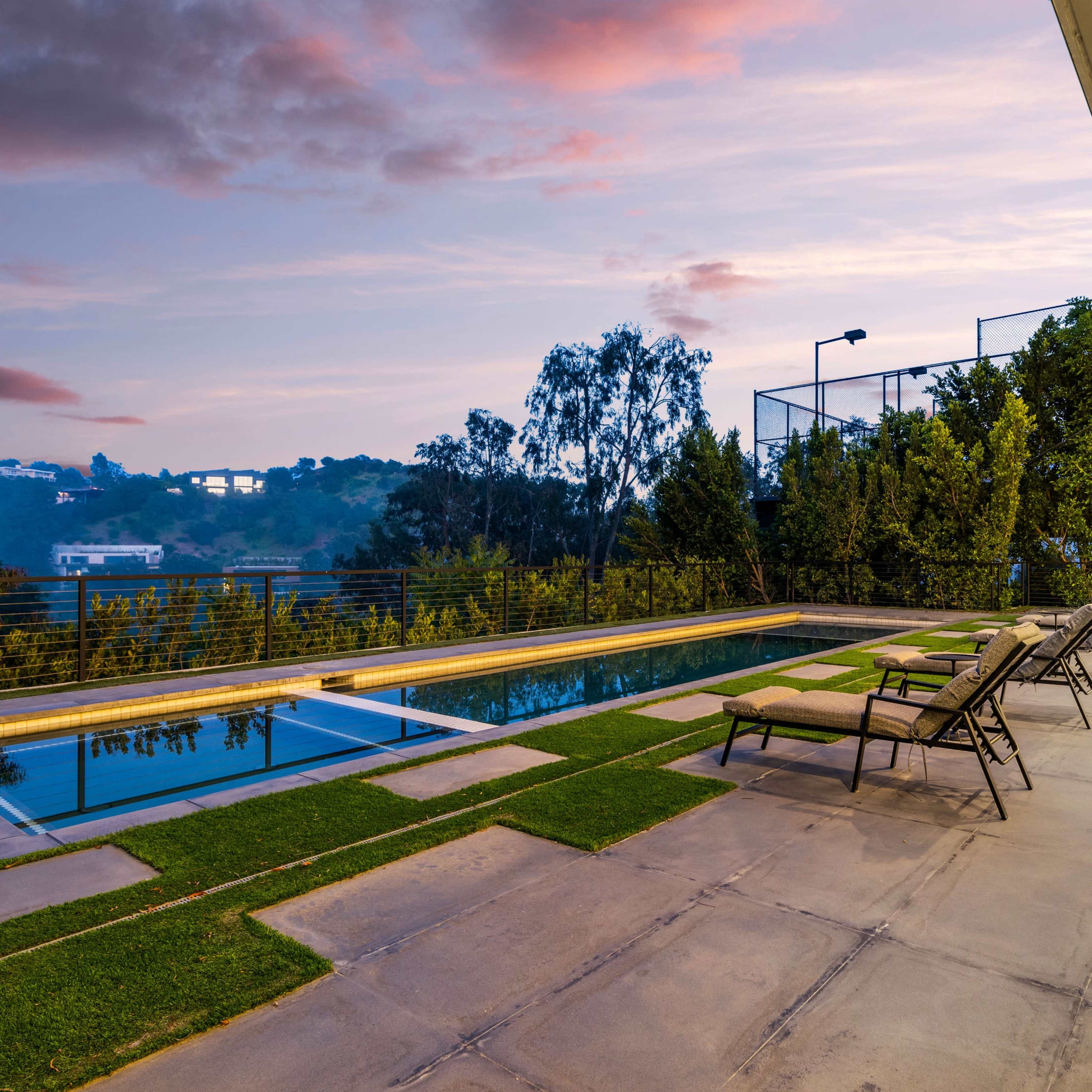 A modern outdoor patio with a swimming pool, lounge chairs, and landscaped greenery against a twilight sky.