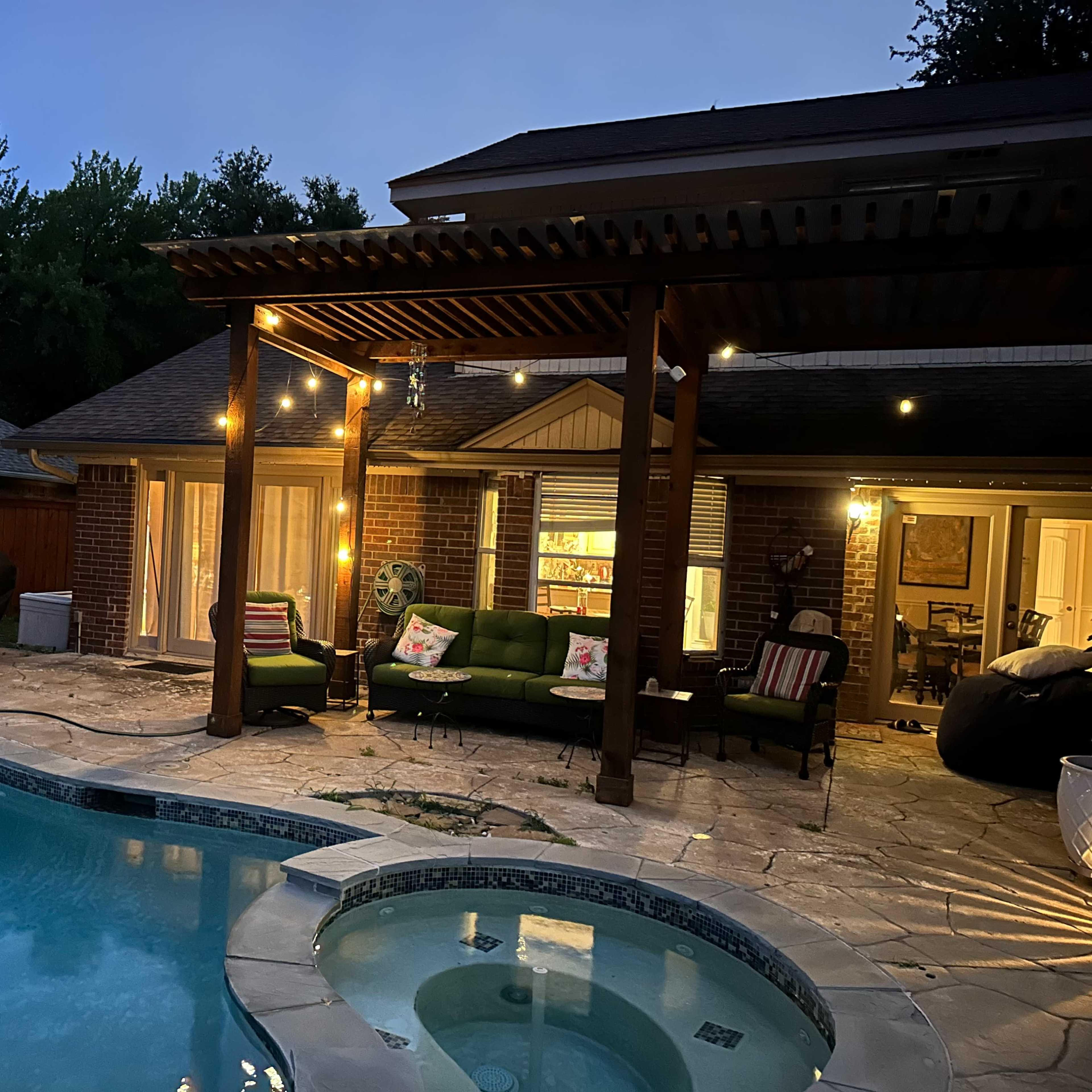 A backyard with a pool, hot tub, patio furniture, and lights strung along a wooden pergola at dusk.