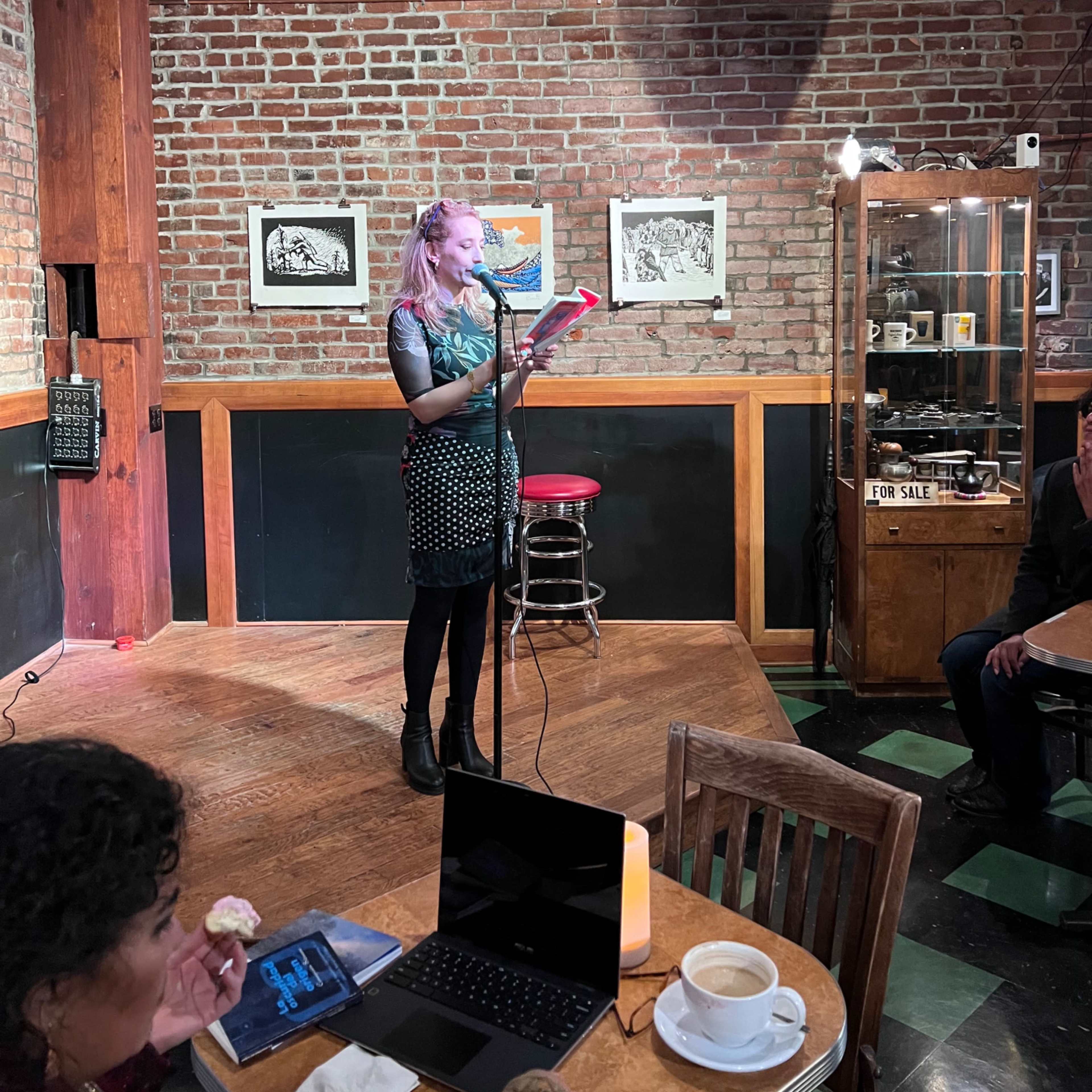 A woman stands at a microphone reading from a book in a brick-walled café with tables and chairs nearby.