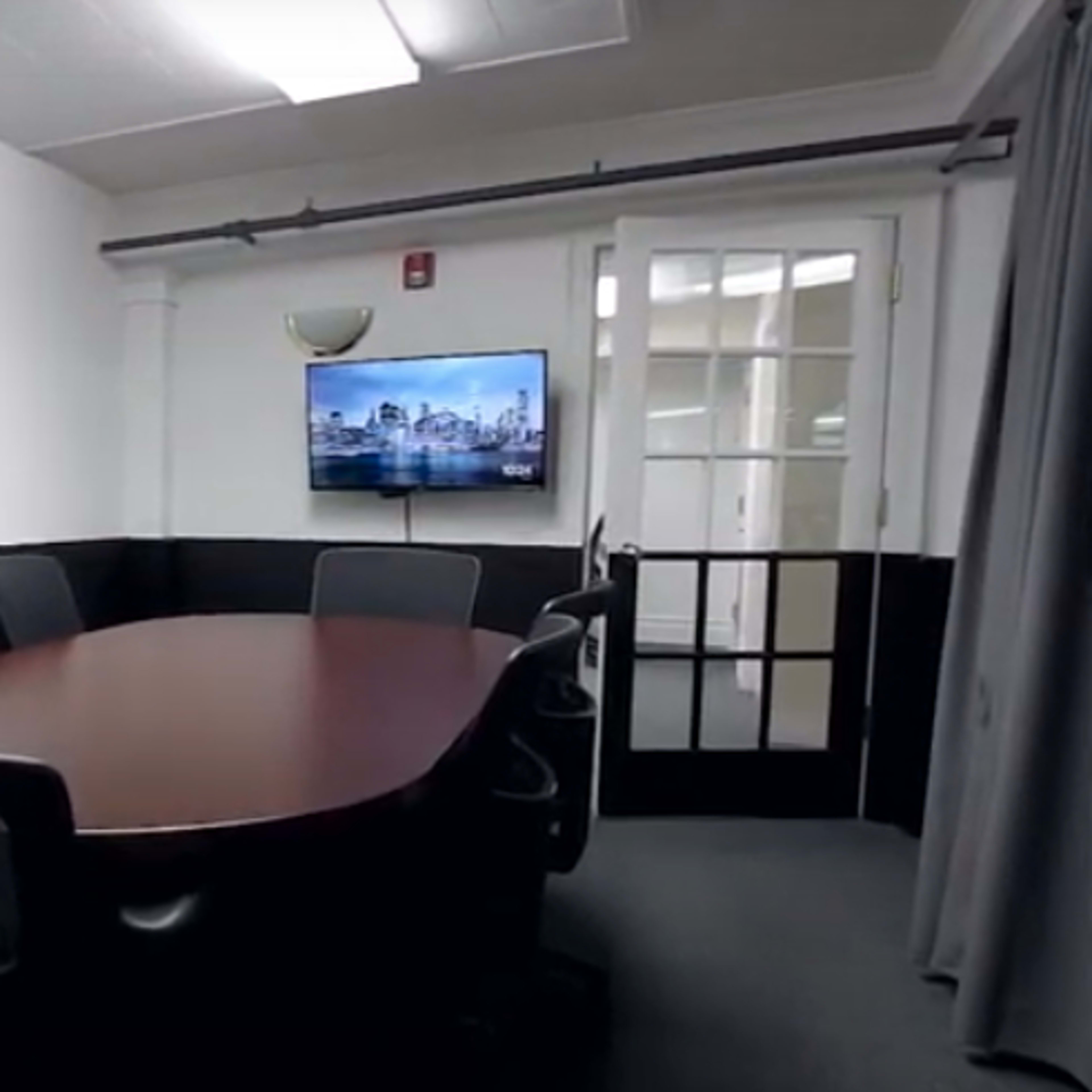 A conference room features a large oval table surrounded by chairs and a mounted television on the wall.