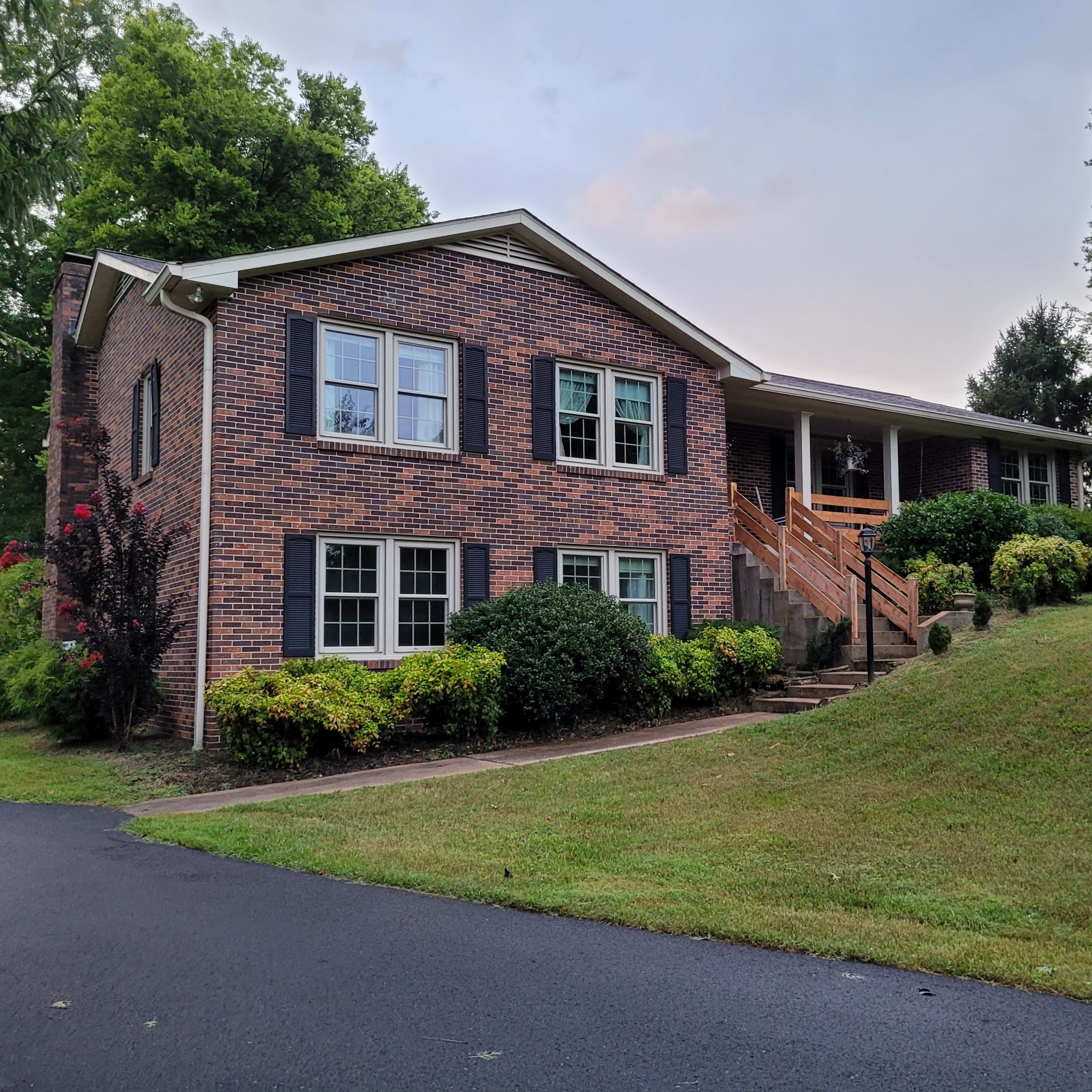 A two-story brick house with blue shutters sits on a grassy slope, accompanied by well-kept shrubbery and a gravel driveway.