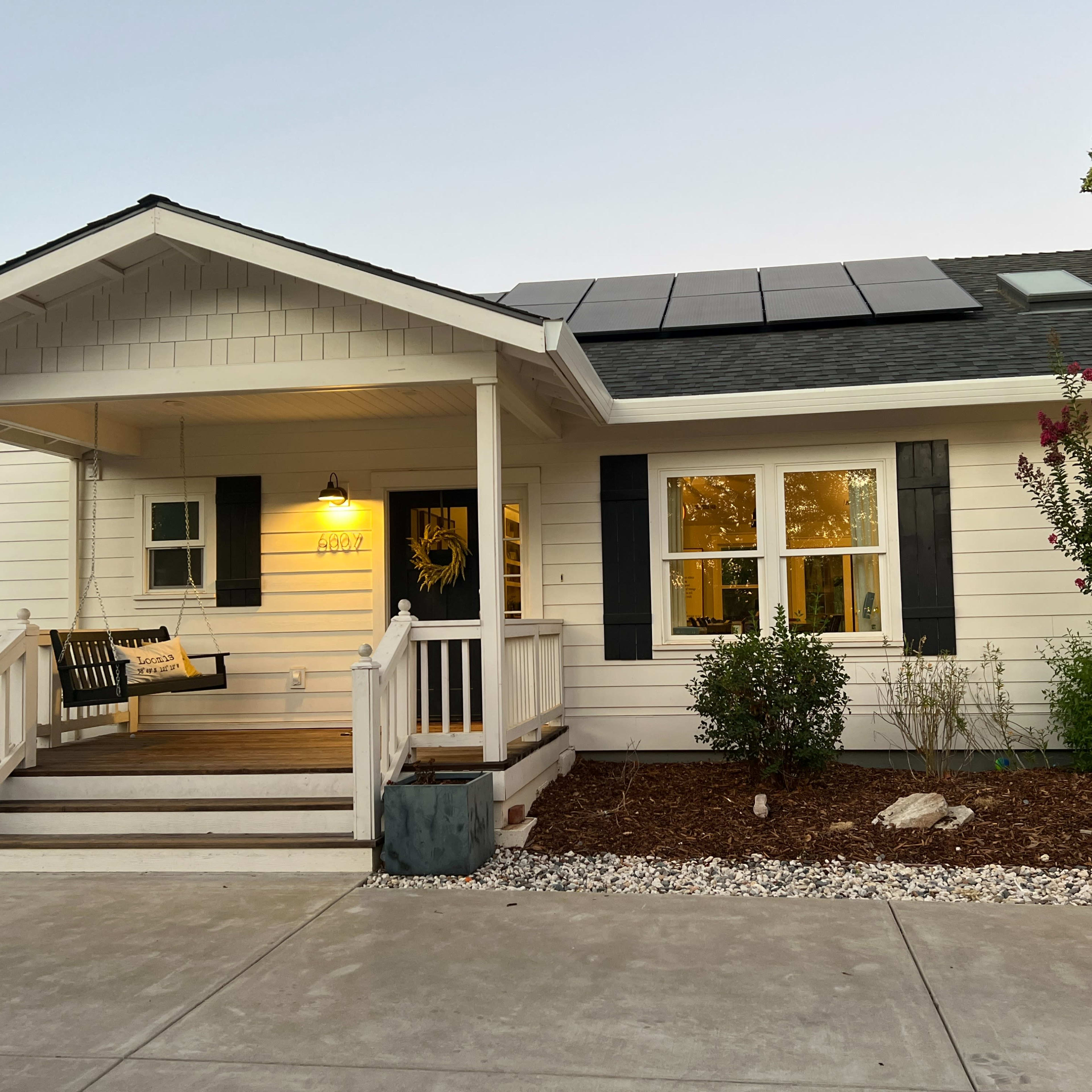 A single-story house features a front porch with a swing, solar panels on the roof, and landscaped gardens.