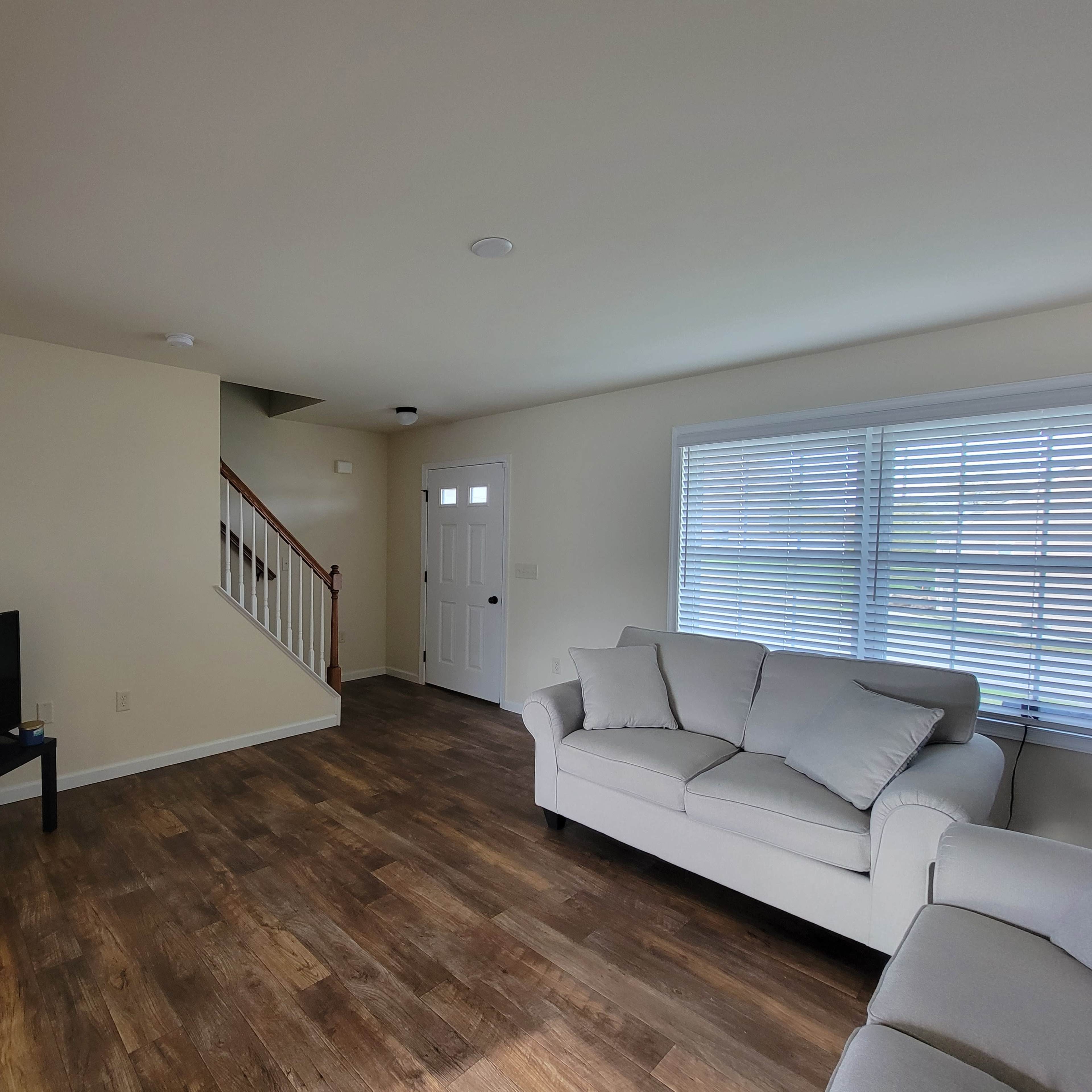 A living room featuring a light-colored sofa, a television on a stand, wooden flooring, and a staircase leading to an upper level.