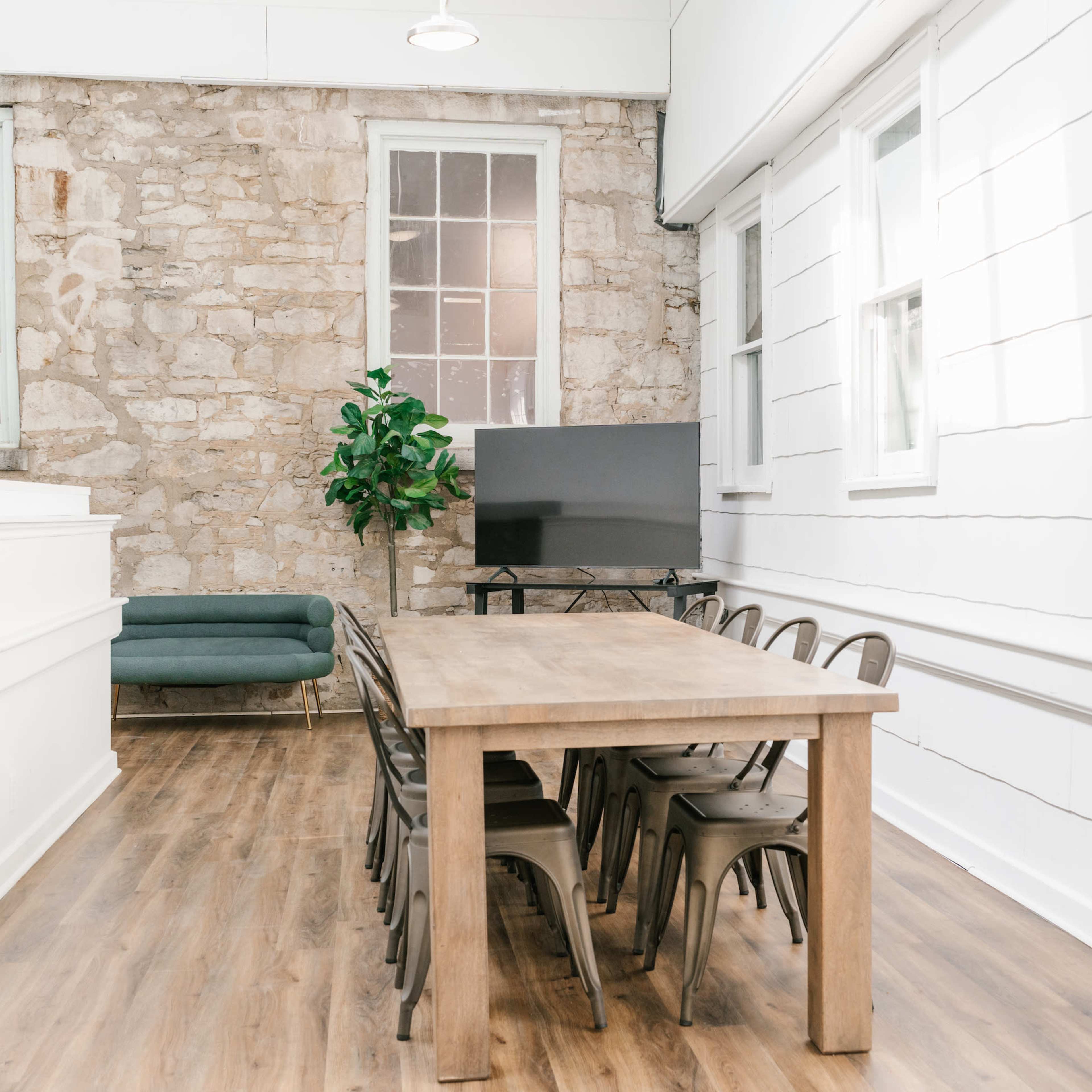 A spacious room featuring a wooden dining table surrounded by metal chairs, with a green sofa and a television against a stone wall.