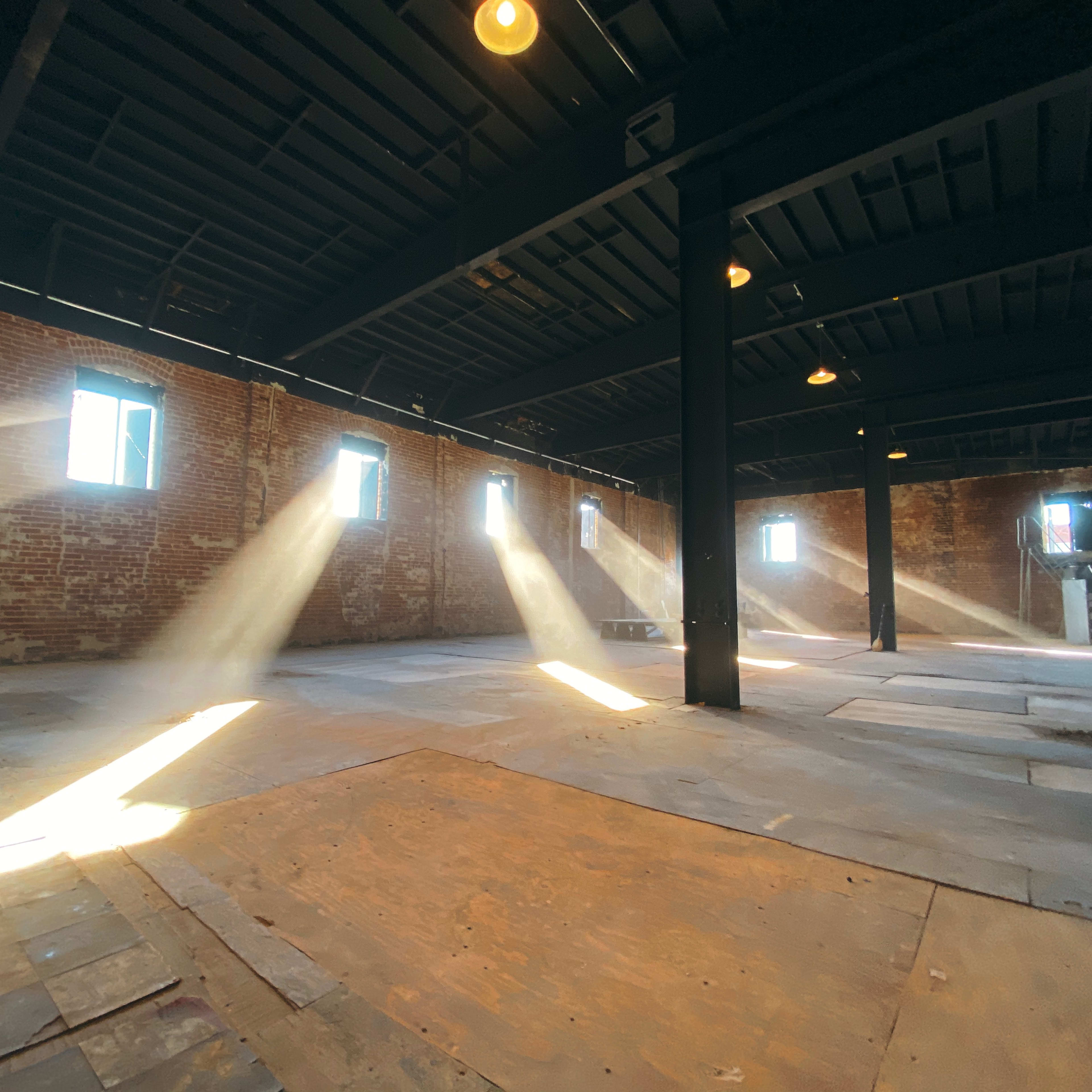 Sunlight streams through windows onto the floor of an empty brick room with a black ceiling and exposed beams.
