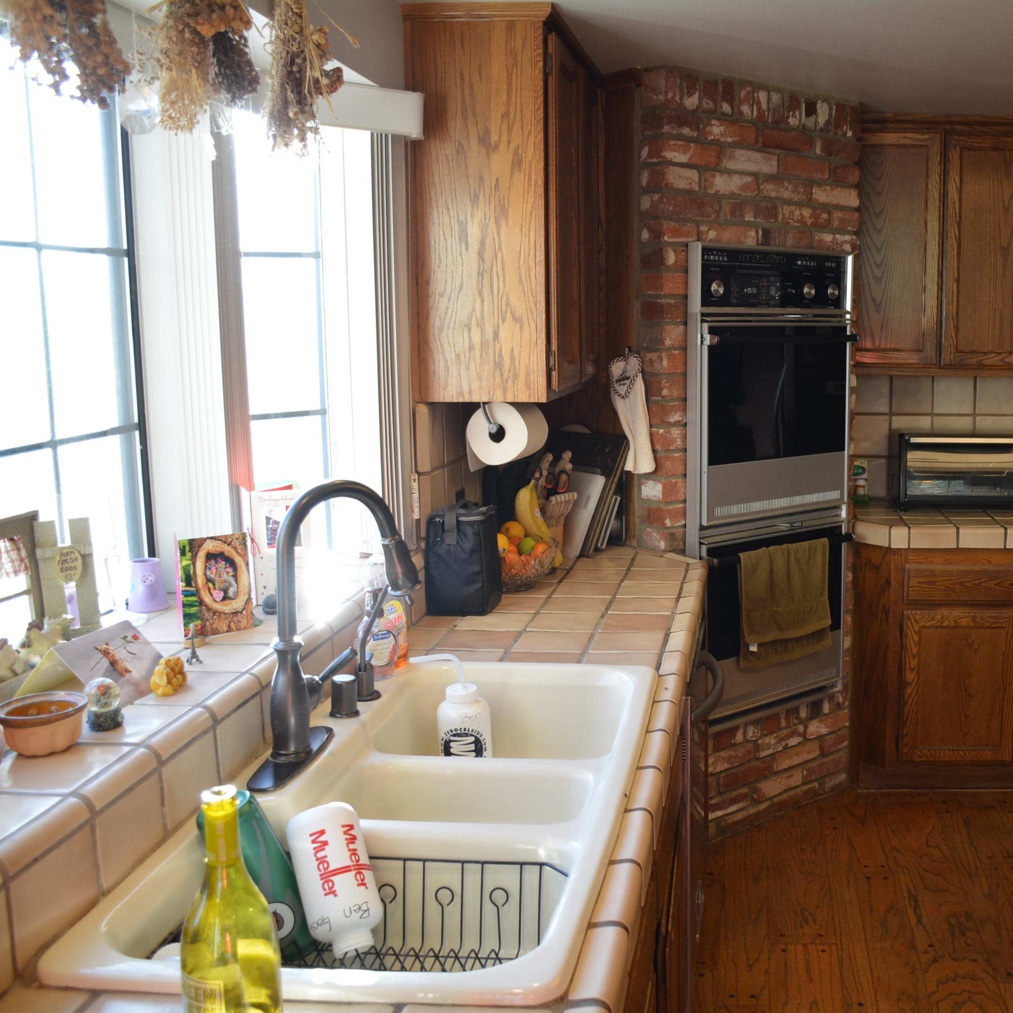 A kitchen with wooden cabinets, a tiled countertop, a double sink, a stove, and an oven, along with various kitchen appliances and decorations.