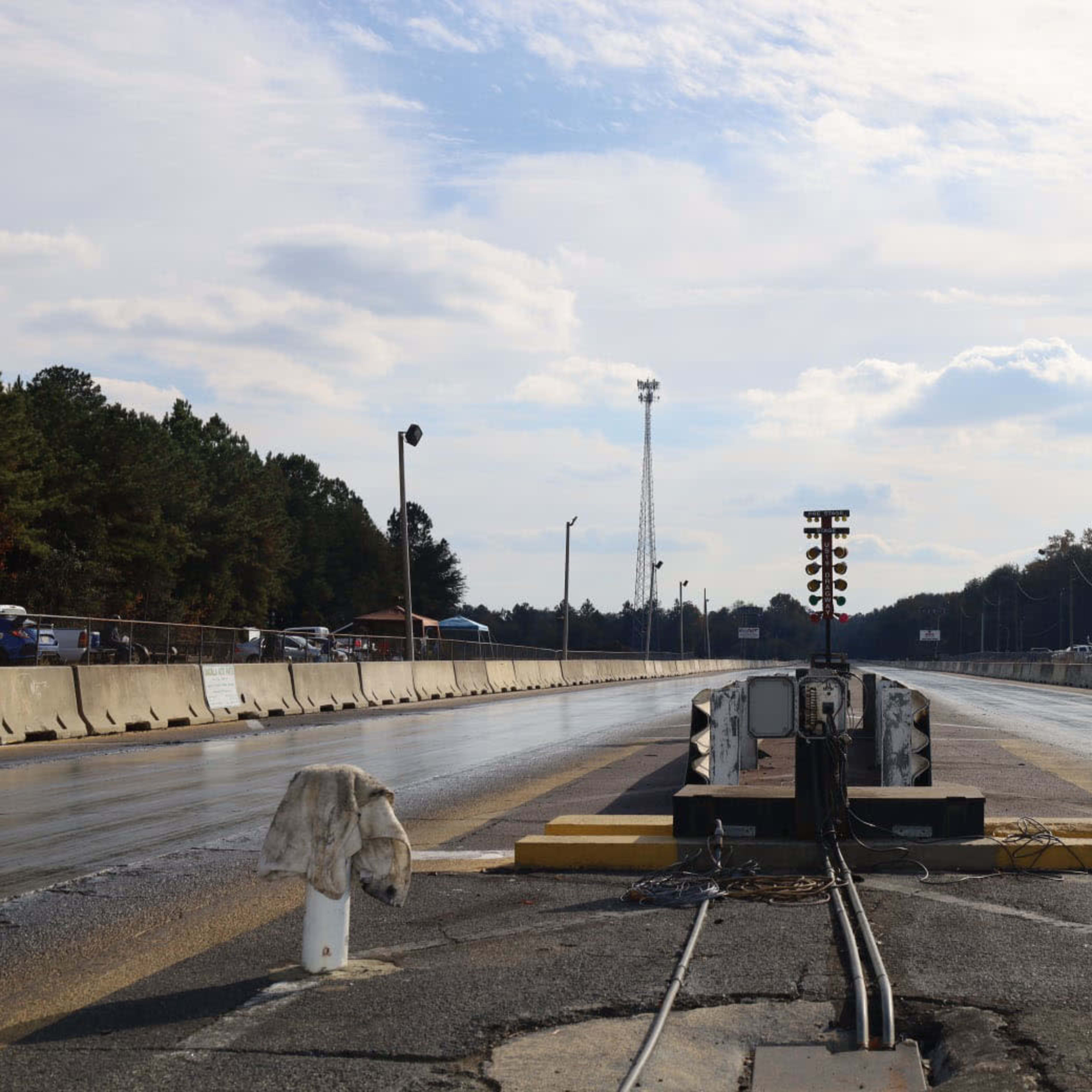 A drag racing track with a starting light system at the end, bordered by concrete barriers and trees in the background under a cloudy sky.
