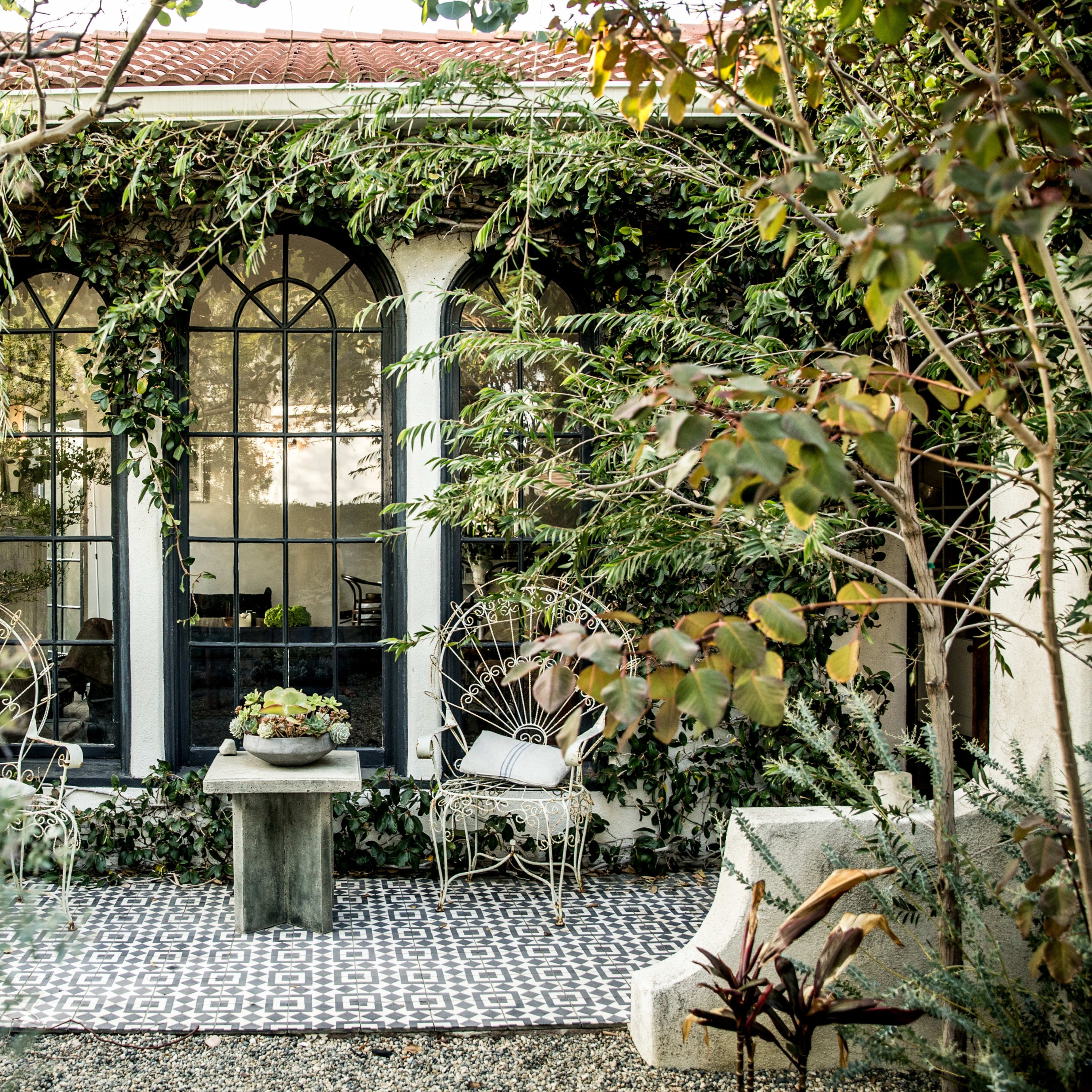 A stone table is surrounded by two wicker chairs on a patterned tile patio, framed by lush greenery and the large windows of a house.