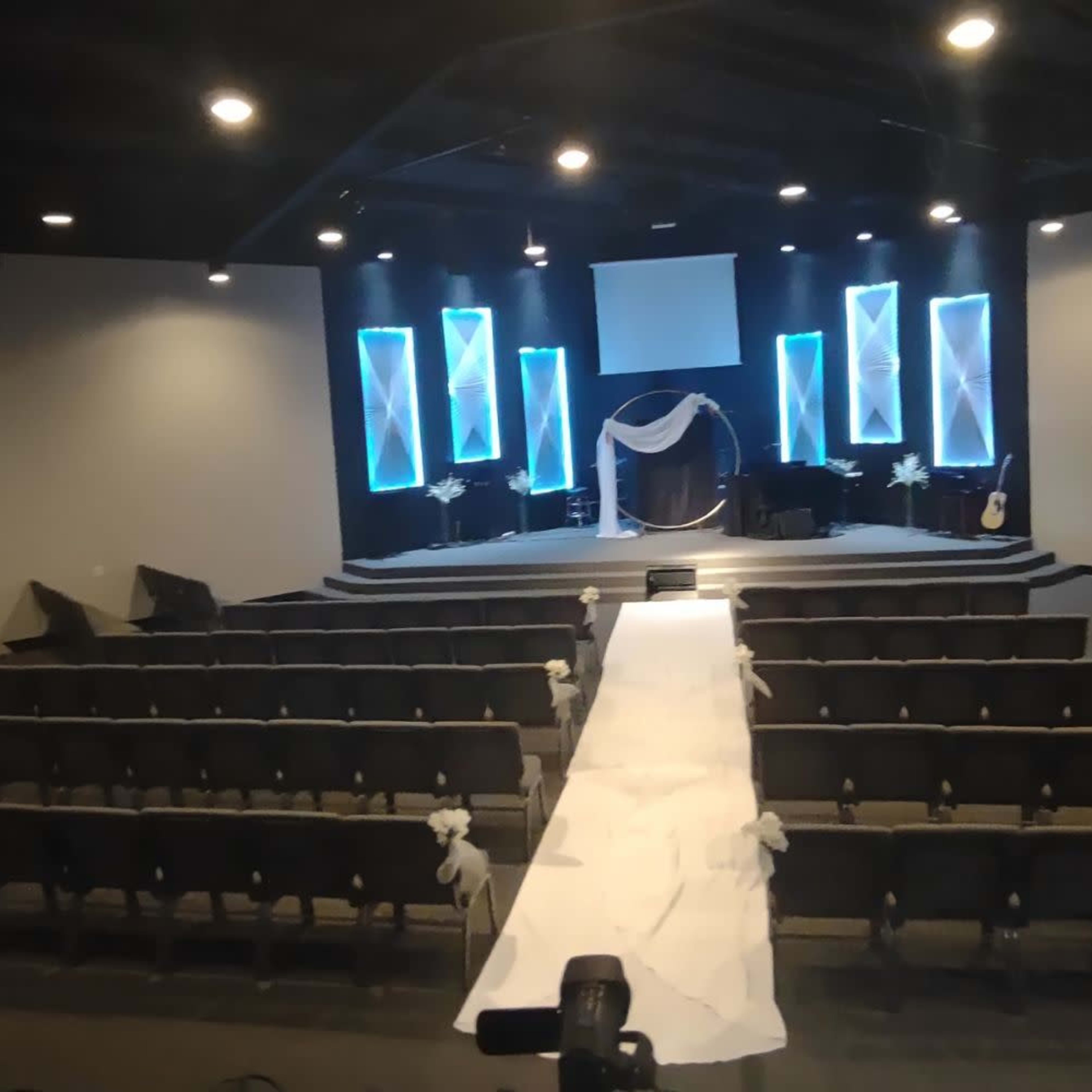 A spacious auditorium set up for an event, featuring rows of chairs facing a stage adorned with blue lighting and decorative elements.