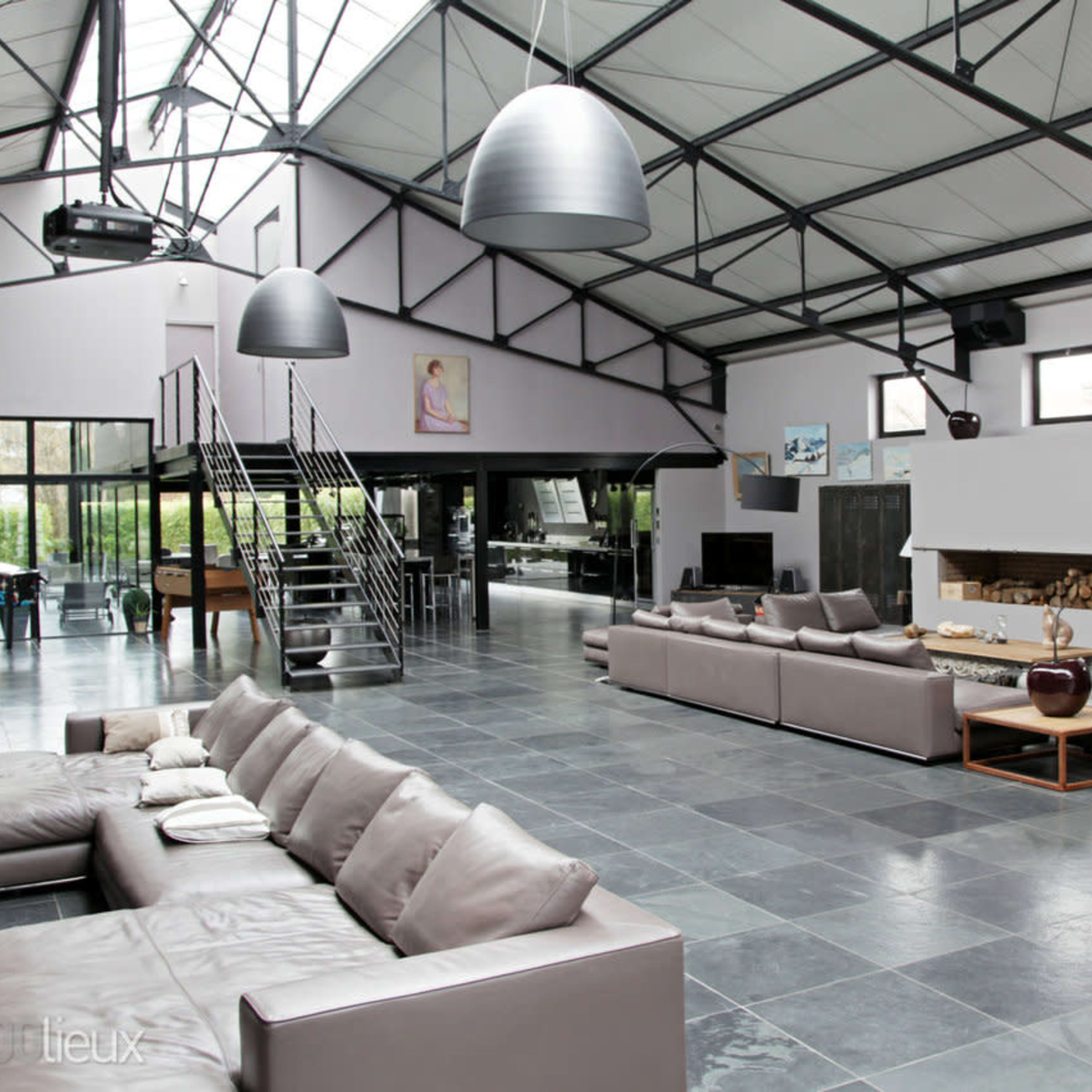 A spacious, modern loft interior featuring leather sofas, high ceilings with metal beams, and large windows allowing natural light.