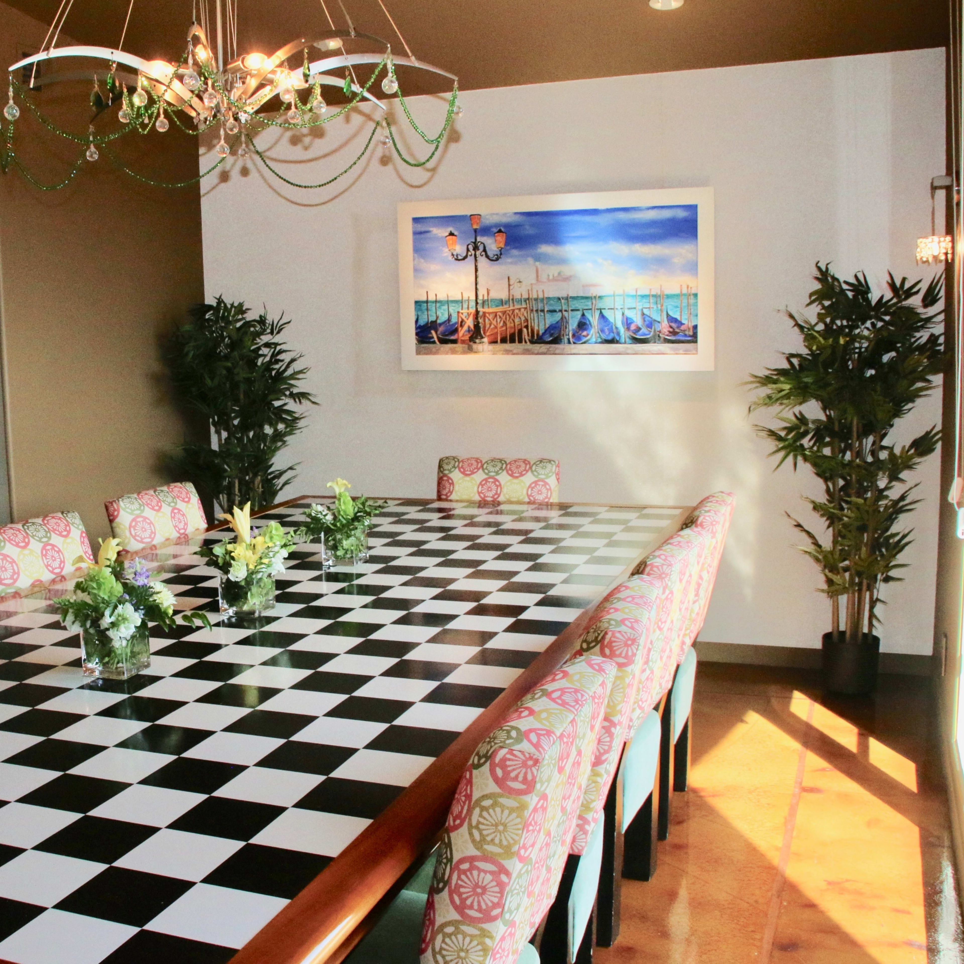 A large dining table with a black and white checkered top is surrounded by chairs featuring floral-patterned cushions, set in a brightly lit room with plants and artwork on the walls.
