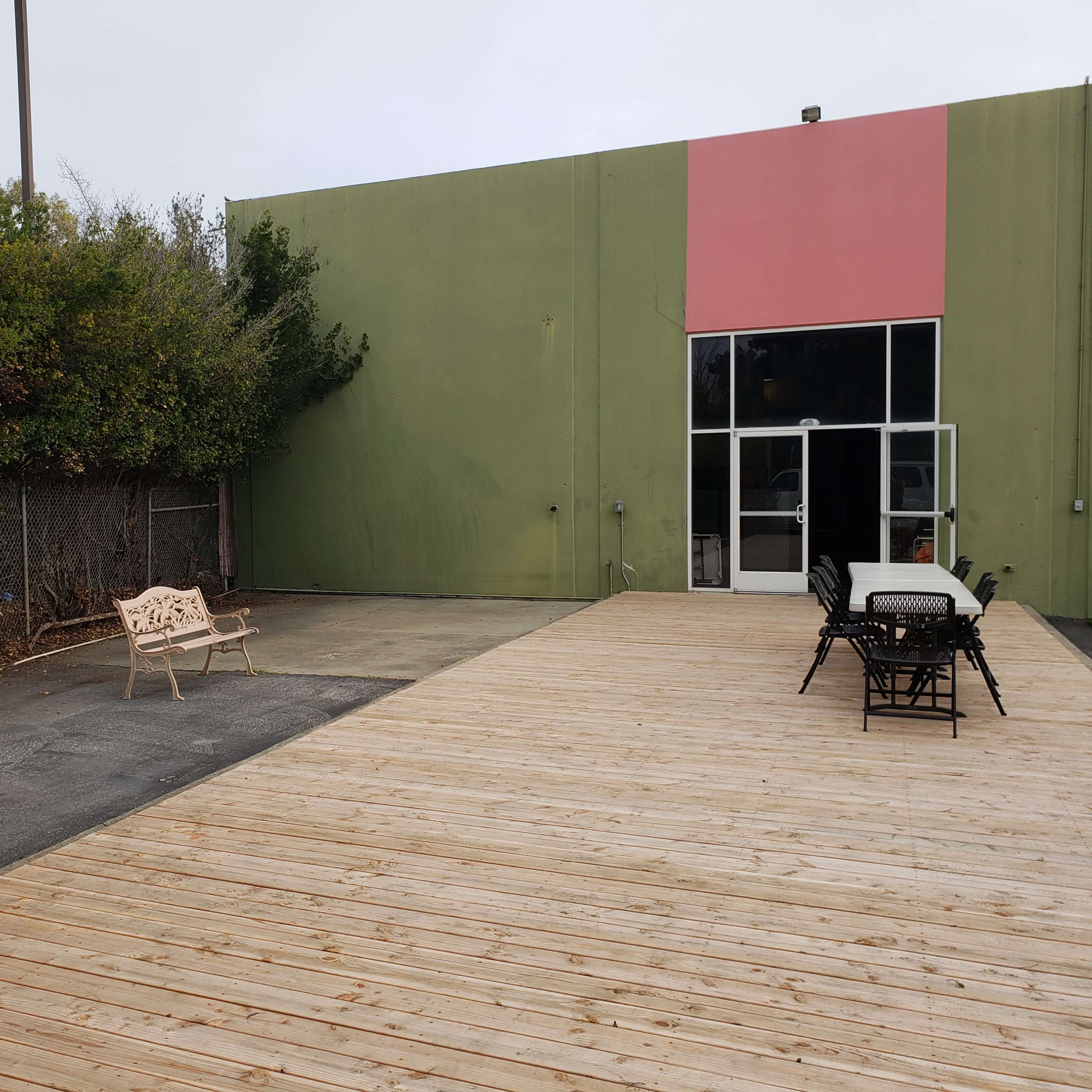 A wooden deck extends from a building with a green wall and a pink section, featuring outdoor seating arrangements and a bench on one side.