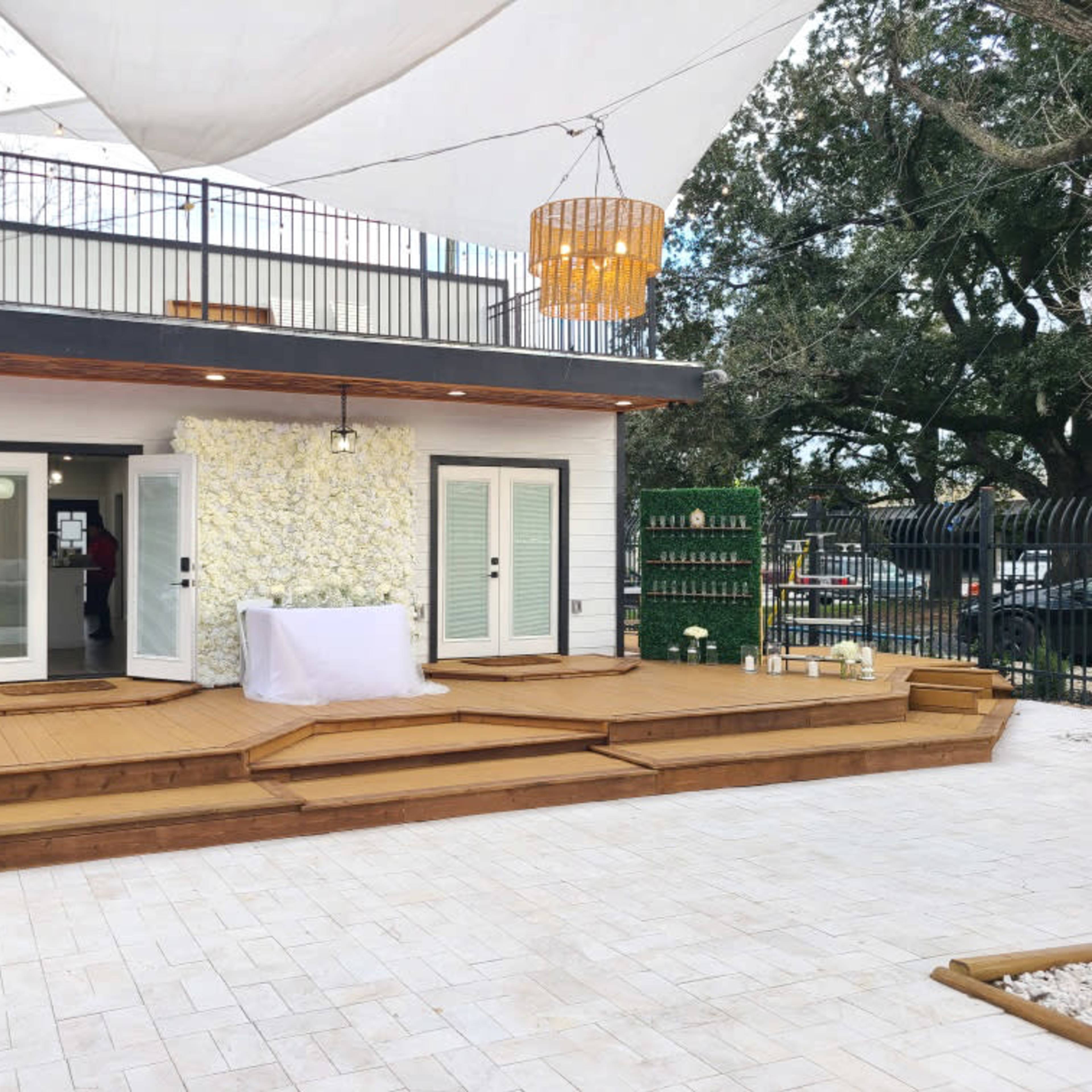 A spacious outdoor deck area with a stage, surrounded by greenery and a modern building featuring large windows and a decorative wall.