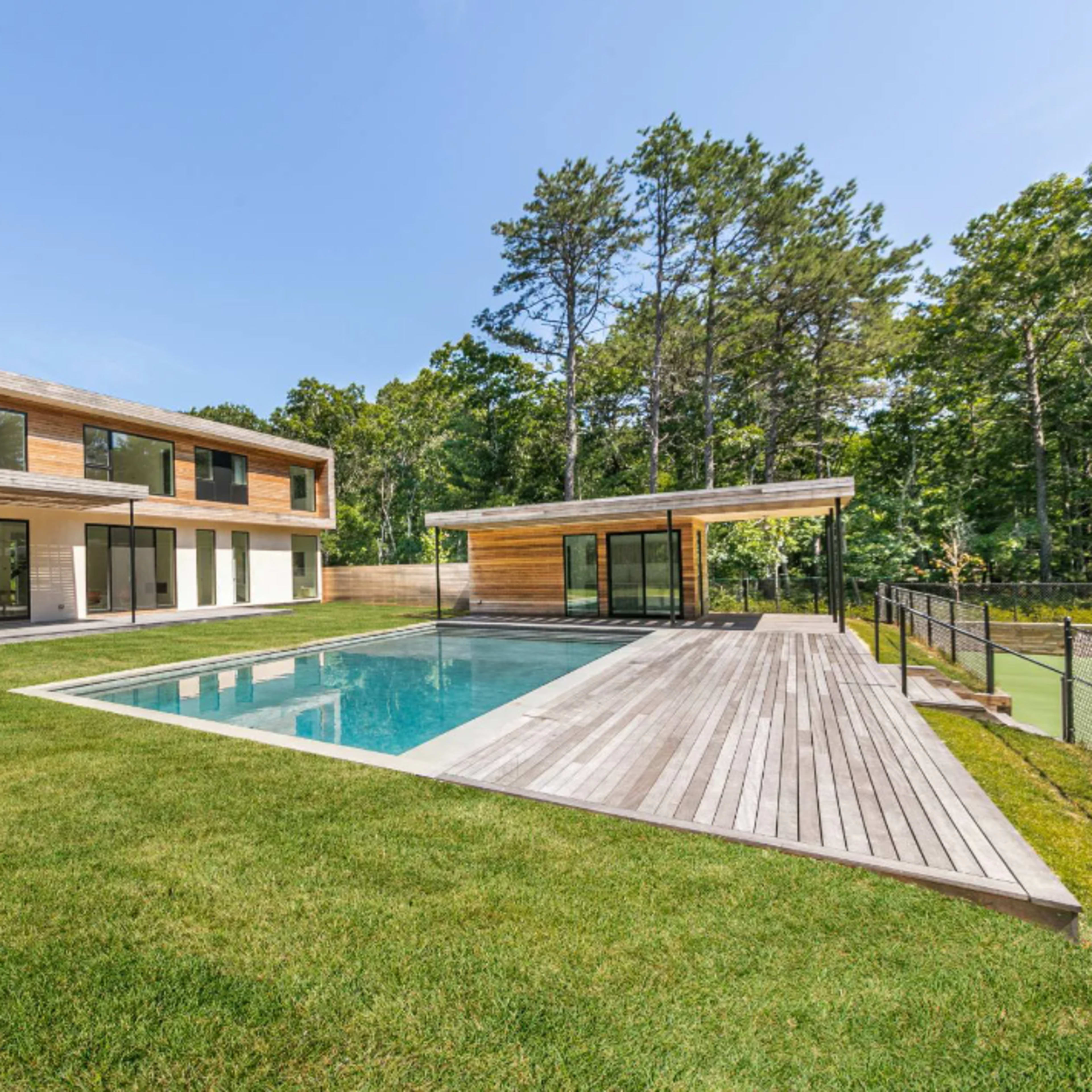 A modern two-story house with large windows and a wooden deck surrounds a rectangular swimming pool in a grassy yard bordered by trees.