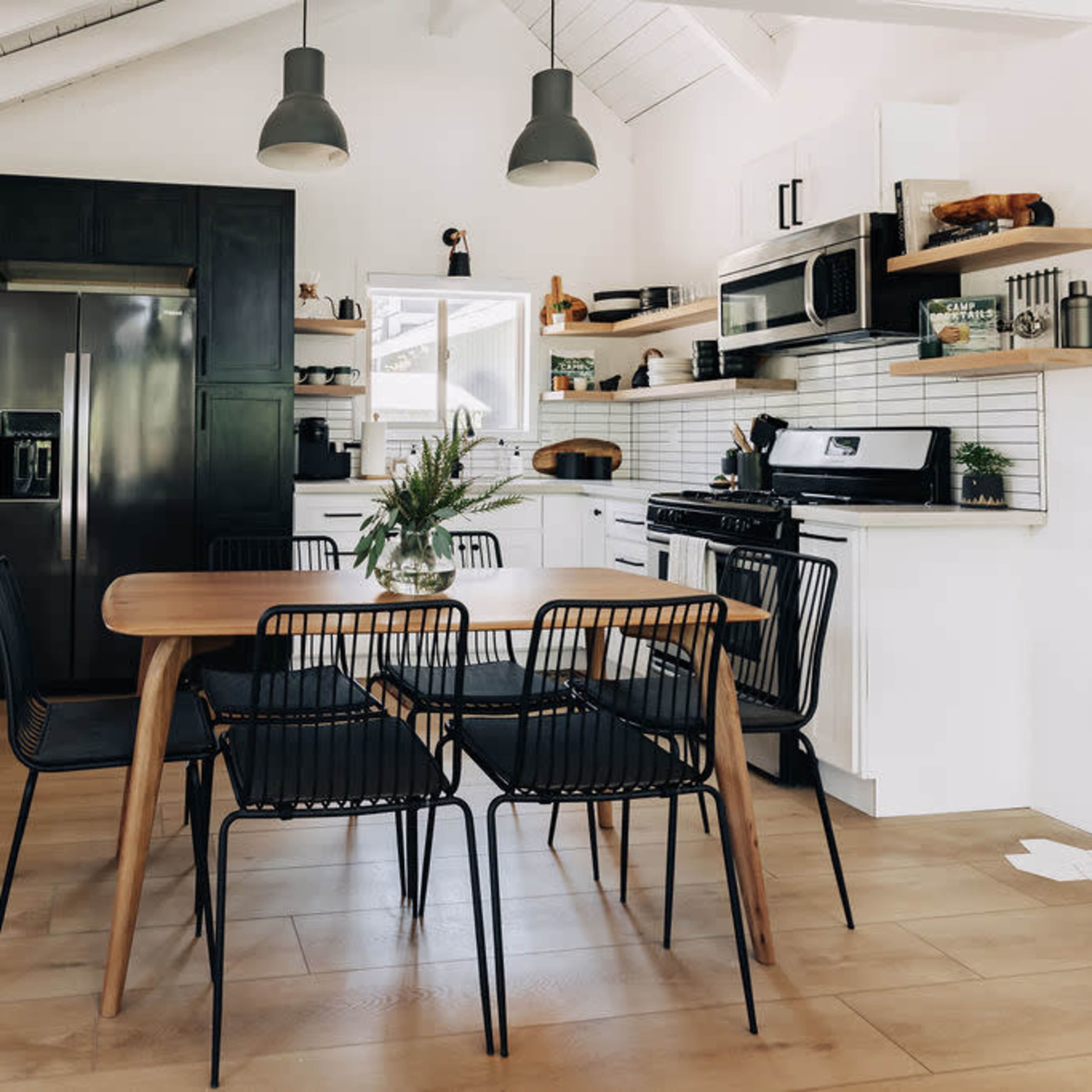 A modern kitchen and dining area features a black table surrounded by six chairs, stainless steel appliances, and a white tile backsplash.