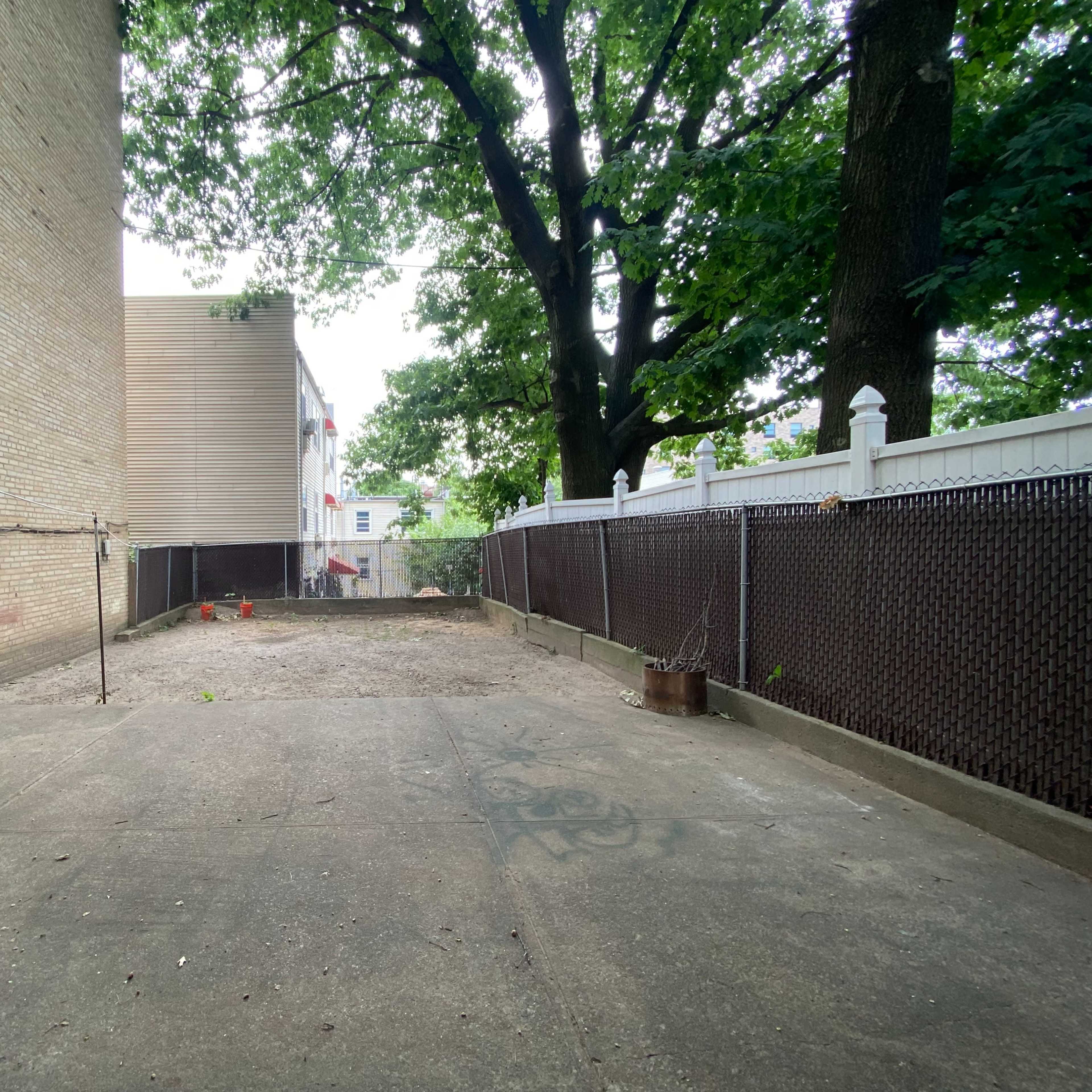 An empty, fenced yard with a concrete surface, surrounded by a brick wall and trees.