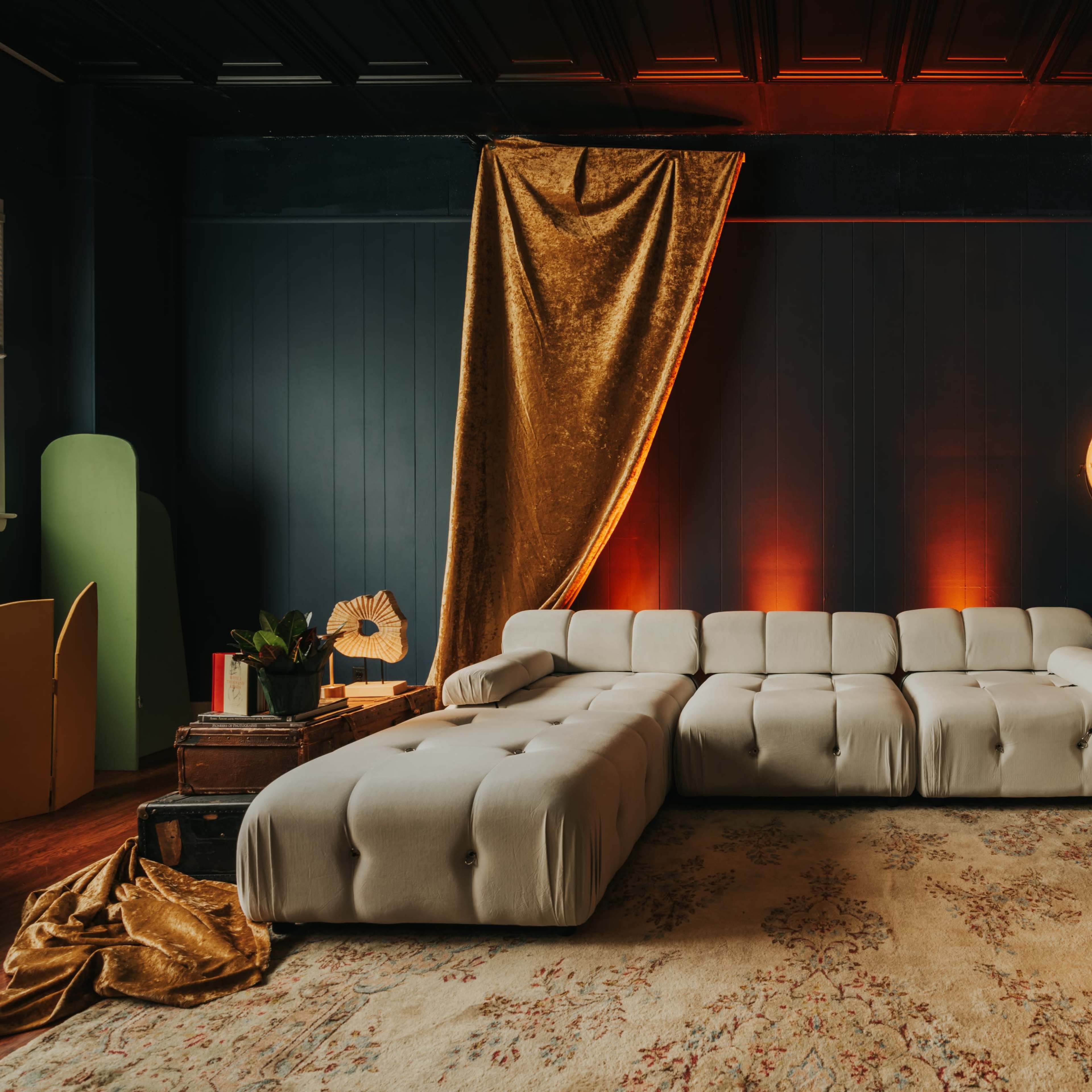 A modern living room featuring a plush sectional sofa, a large area rug, and warm lighting against a dark wall, complemented by decorative plants and a golden drapery.