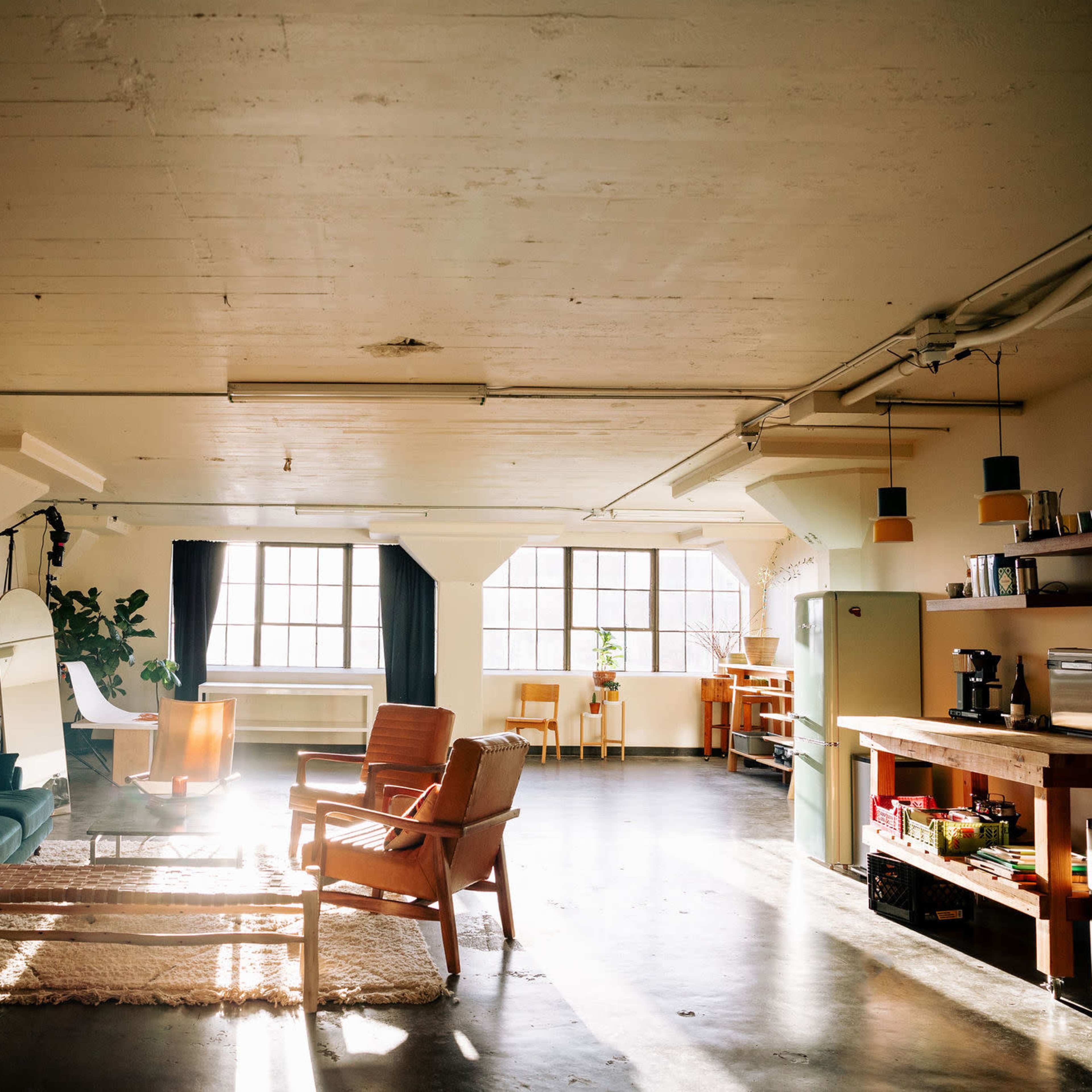 A spacious, bright loft-style room with large windows, featuring a seating area with green and brown chairs, a wooden coffee table, and an open kitchen space.