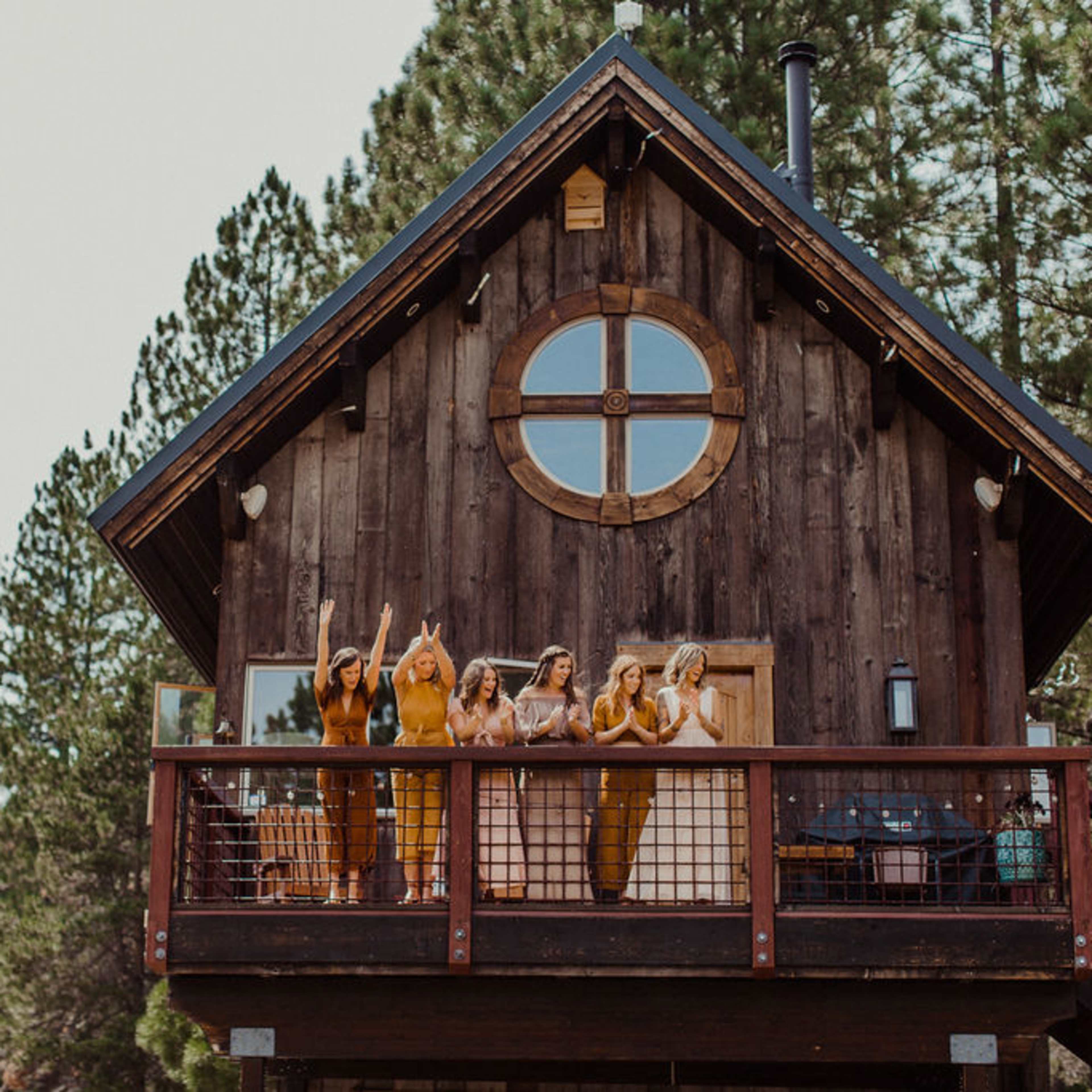 A group of women in matching outfits cheer from the balcony of a wooden cabin surrounded by trees.