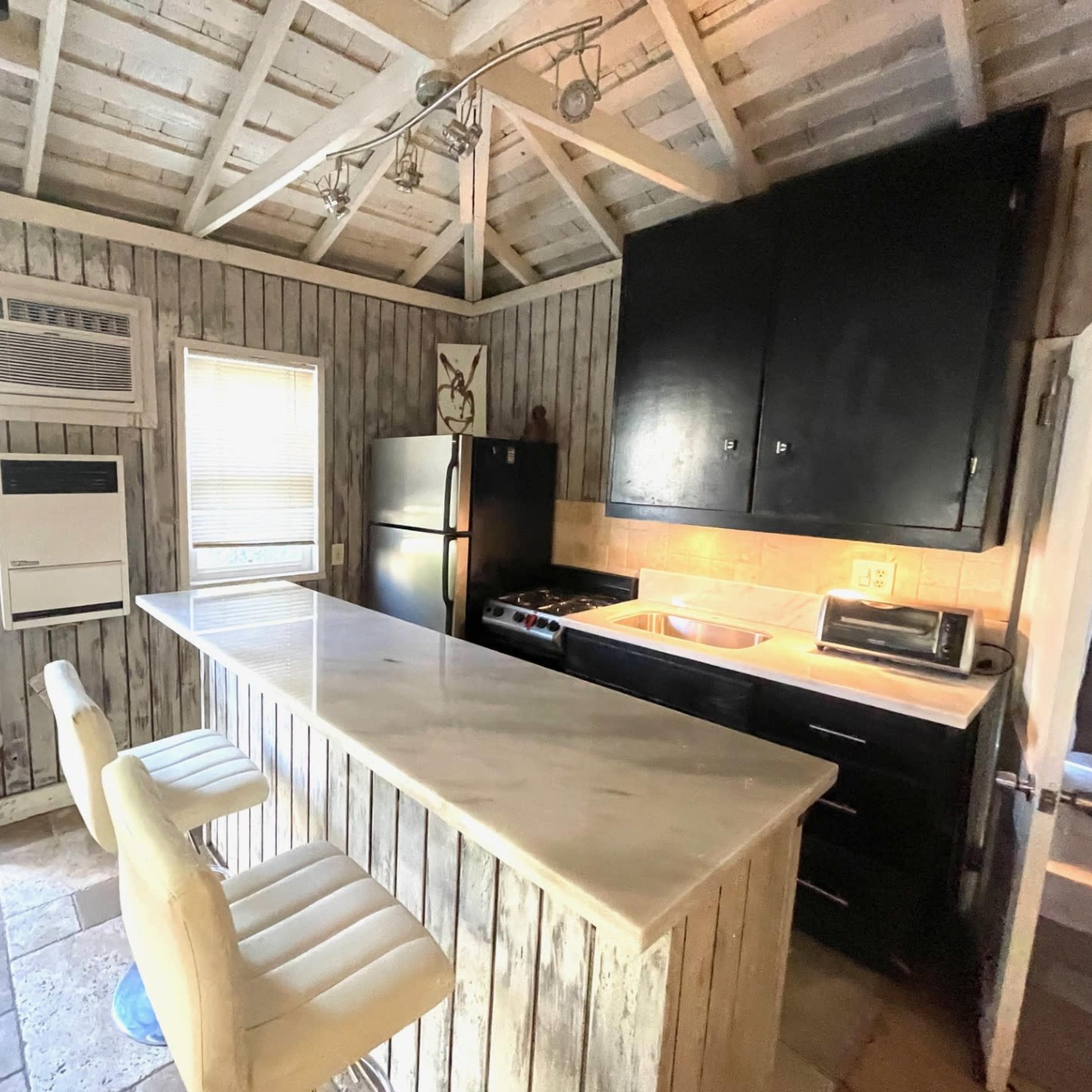 A rustic kitchen with wooden walls, a marble-topped island with two white bar stools, and modern appliances including a refrigerator and a stove.