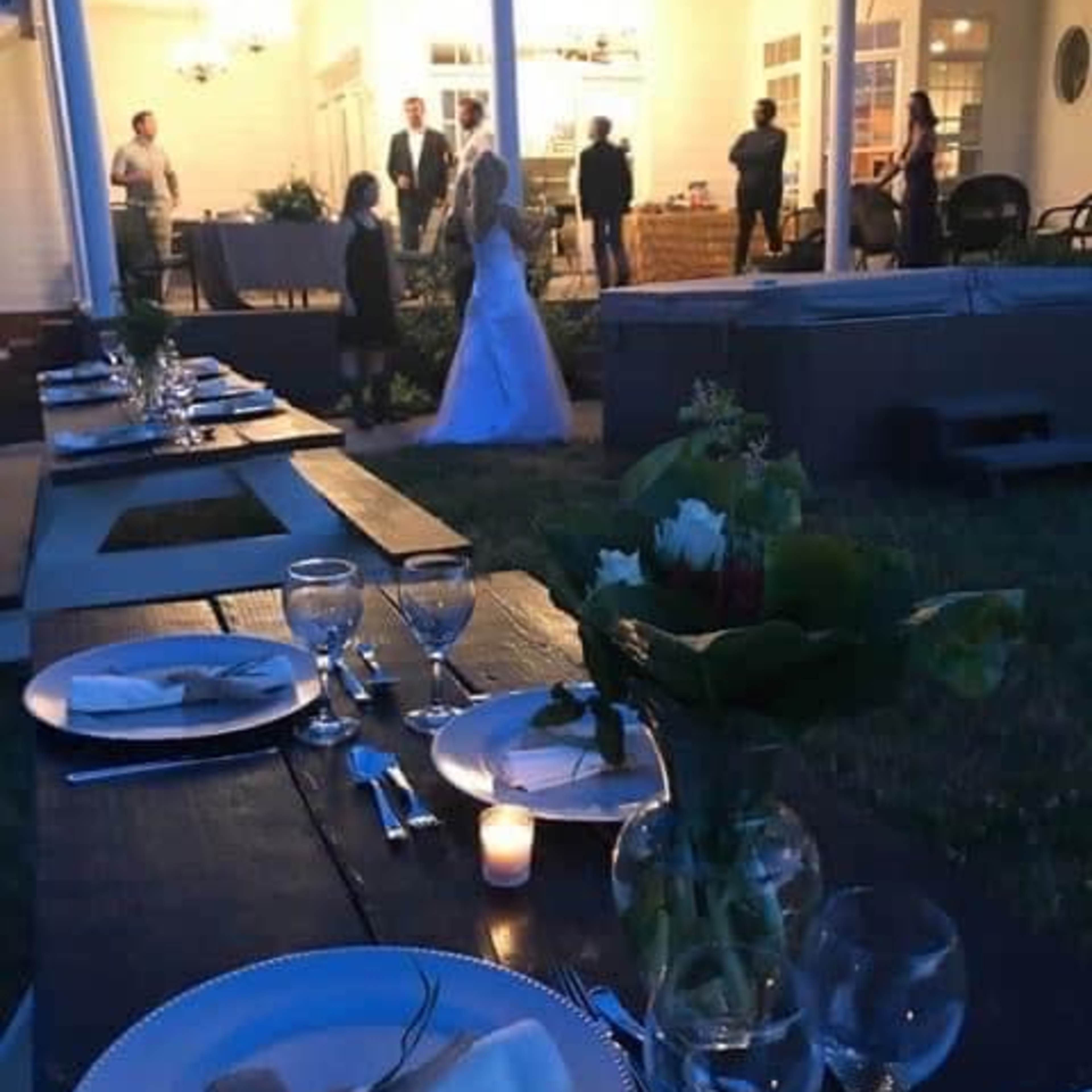 A beautifully arranged outdoor dining table is set with plates and glasses, while a wedding ceremony takes place in the background.