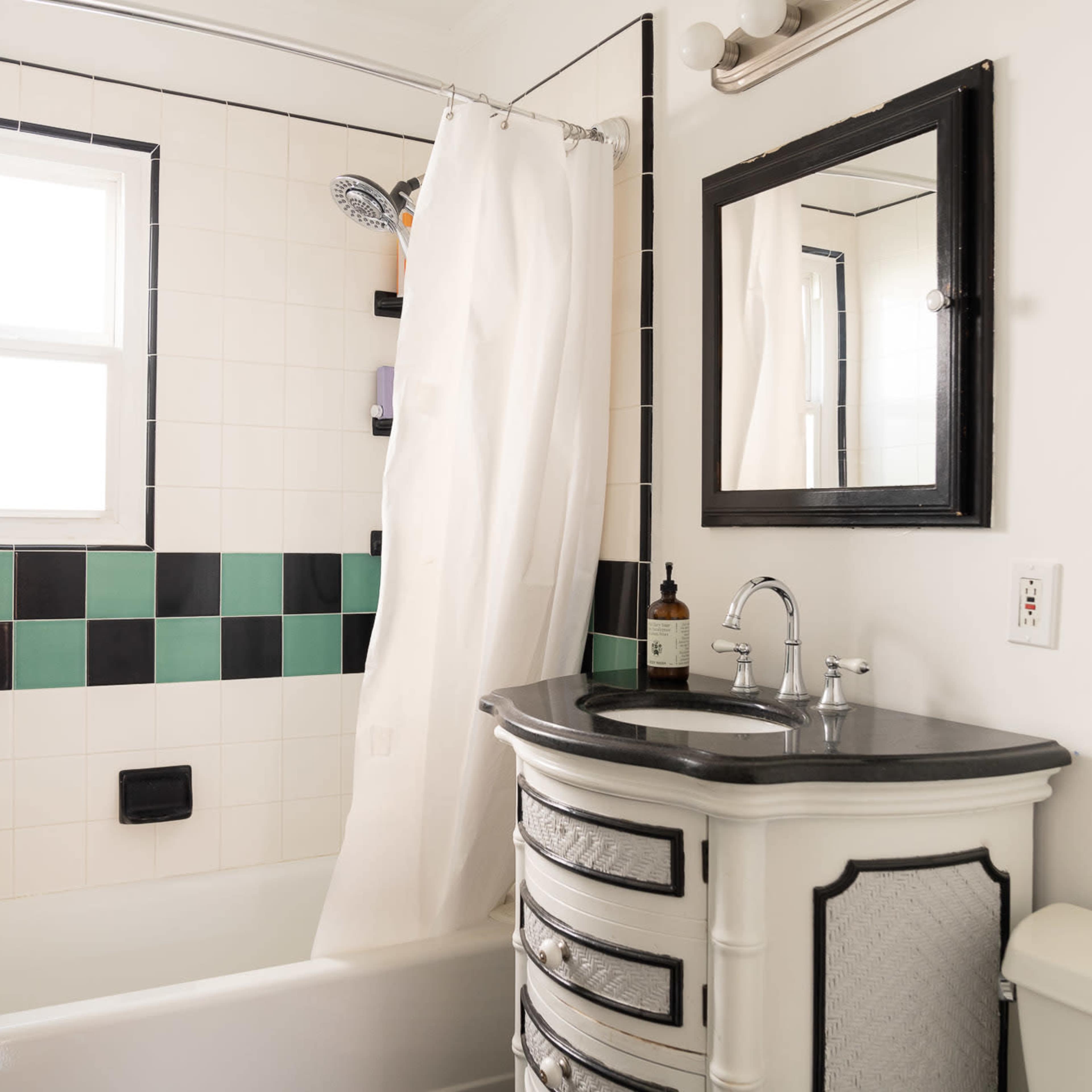 A small bathroom with a tiled bathtub, black and green wall tiles, a shower curtain, and a vanity with a dark countertop and drawers.