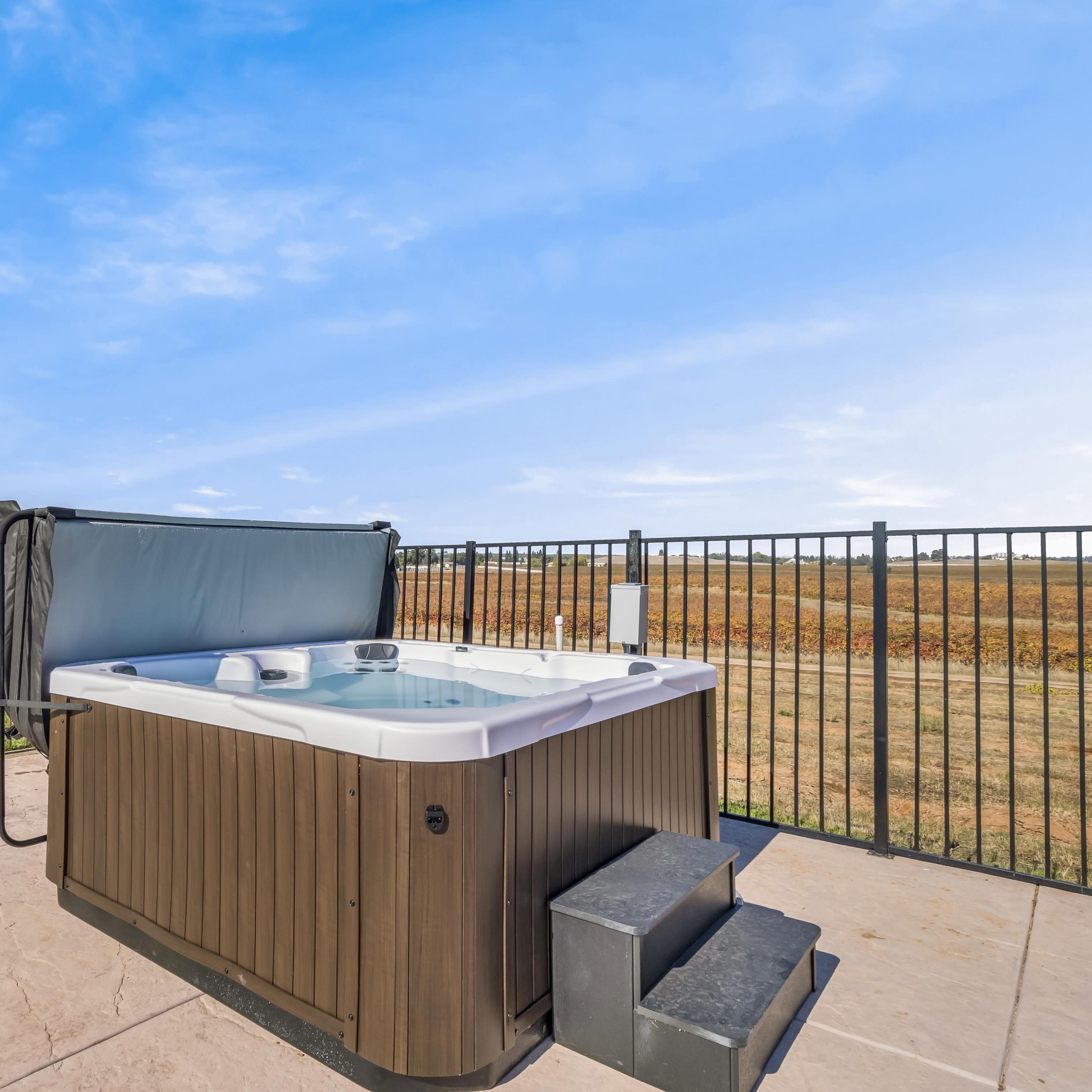 A hot tub is set on a patio surrounded by a fenced yard with a grassy area and a distant view of fields.