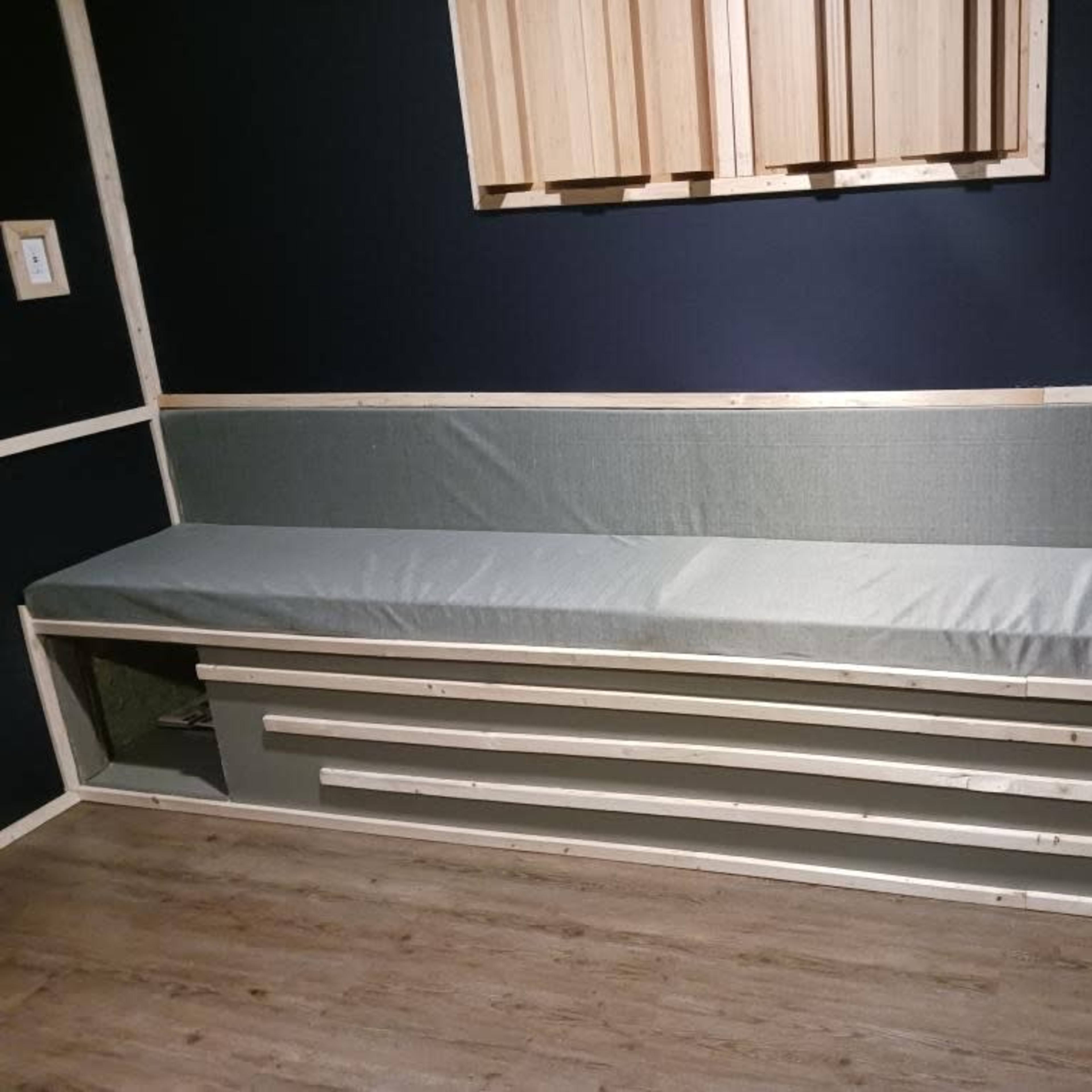 A long, upholstered bench with a wooden frame and storage underneath, set against a dark wall.