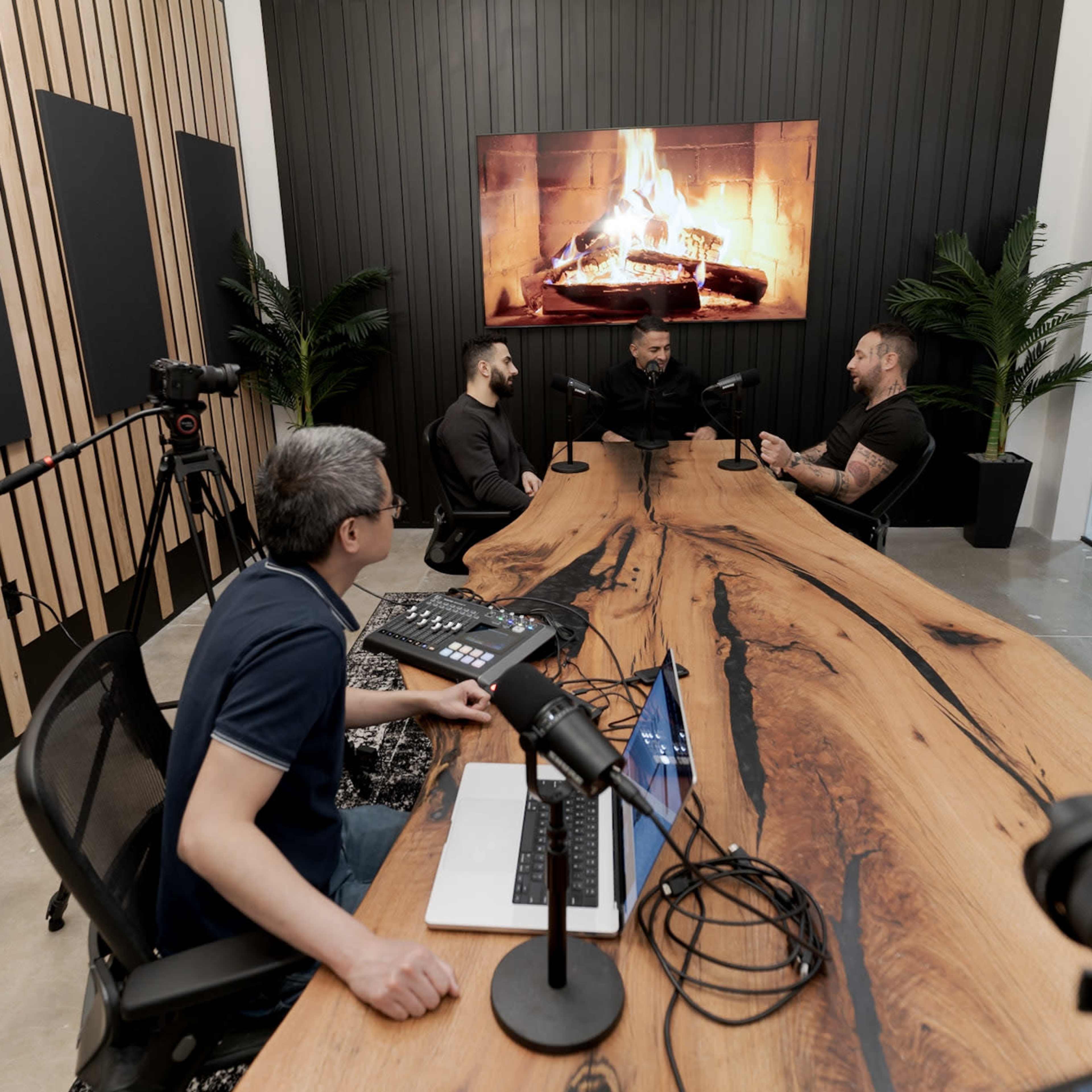 A group of four men engages in discussion around a large wooden table in a modern conference room, with a fireplace video displayed on the wall.