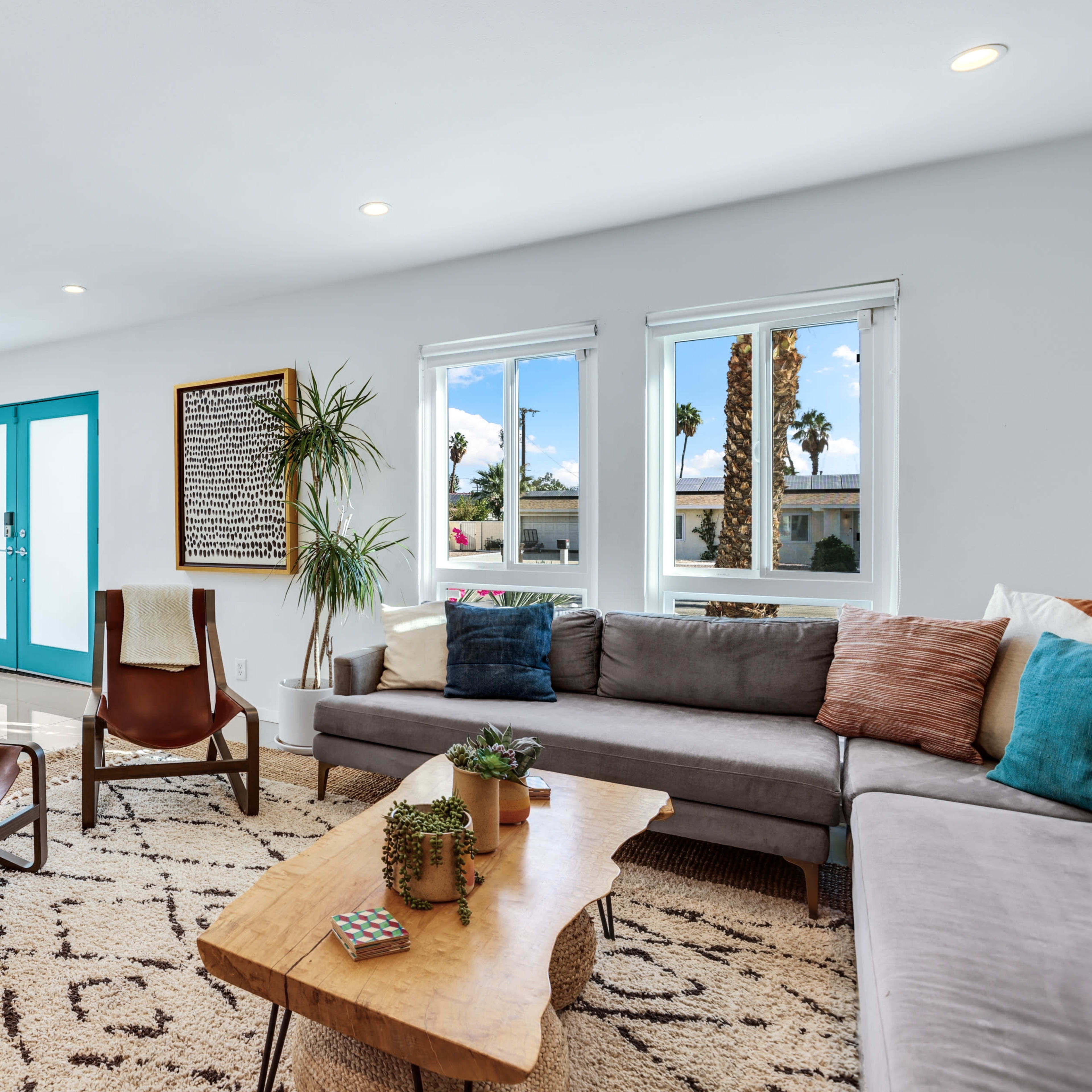 A bright living room with a gray sectional sofa, two wooden chairs, a large patterned rug, and a wooden coffee table, featuring a double door entrance with teal accents and windows that allow natural light in.
