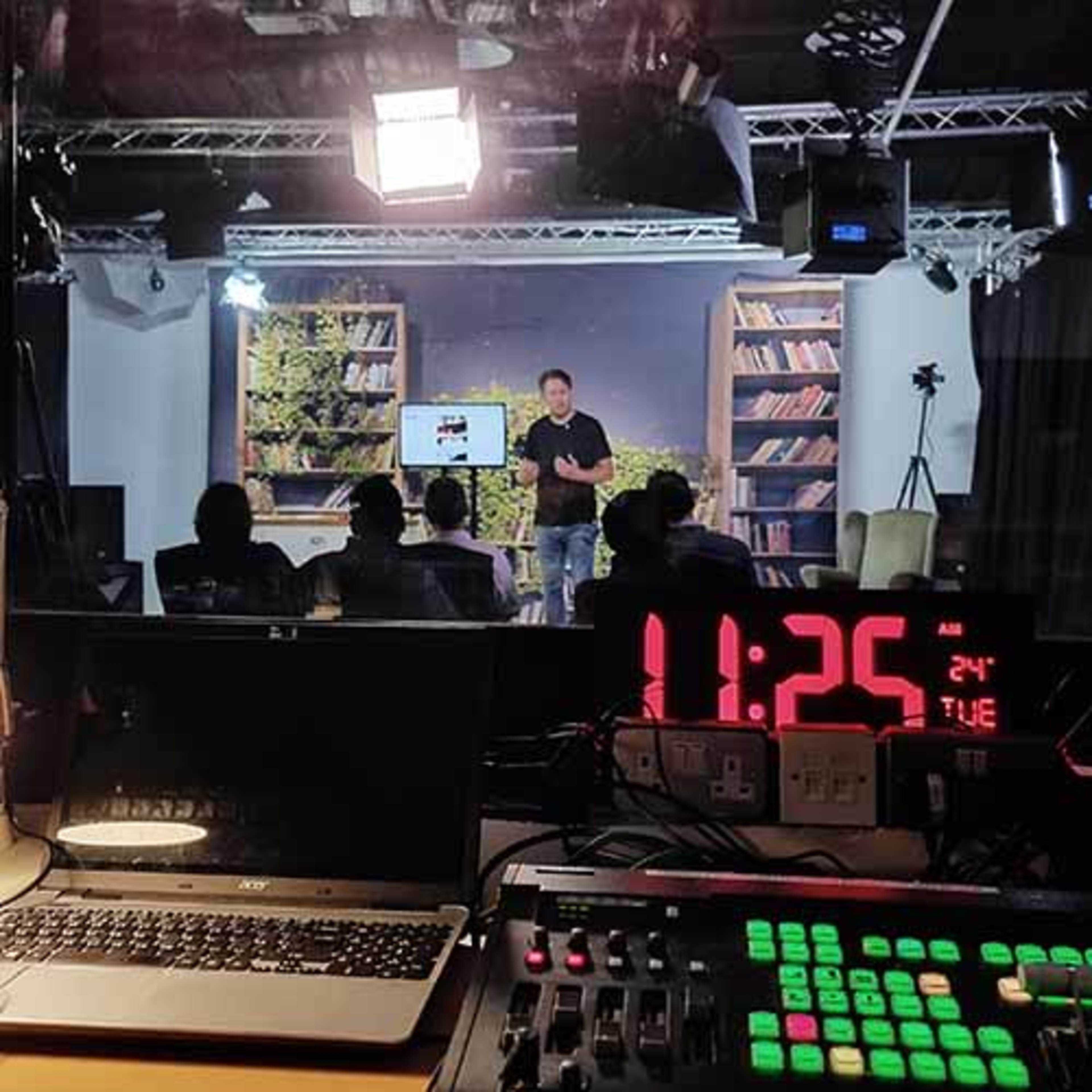 A speaker presents in a studio with a large screen and a bookshelf backdrop, while the audience sits in front and equipment is visible in the foreground.
