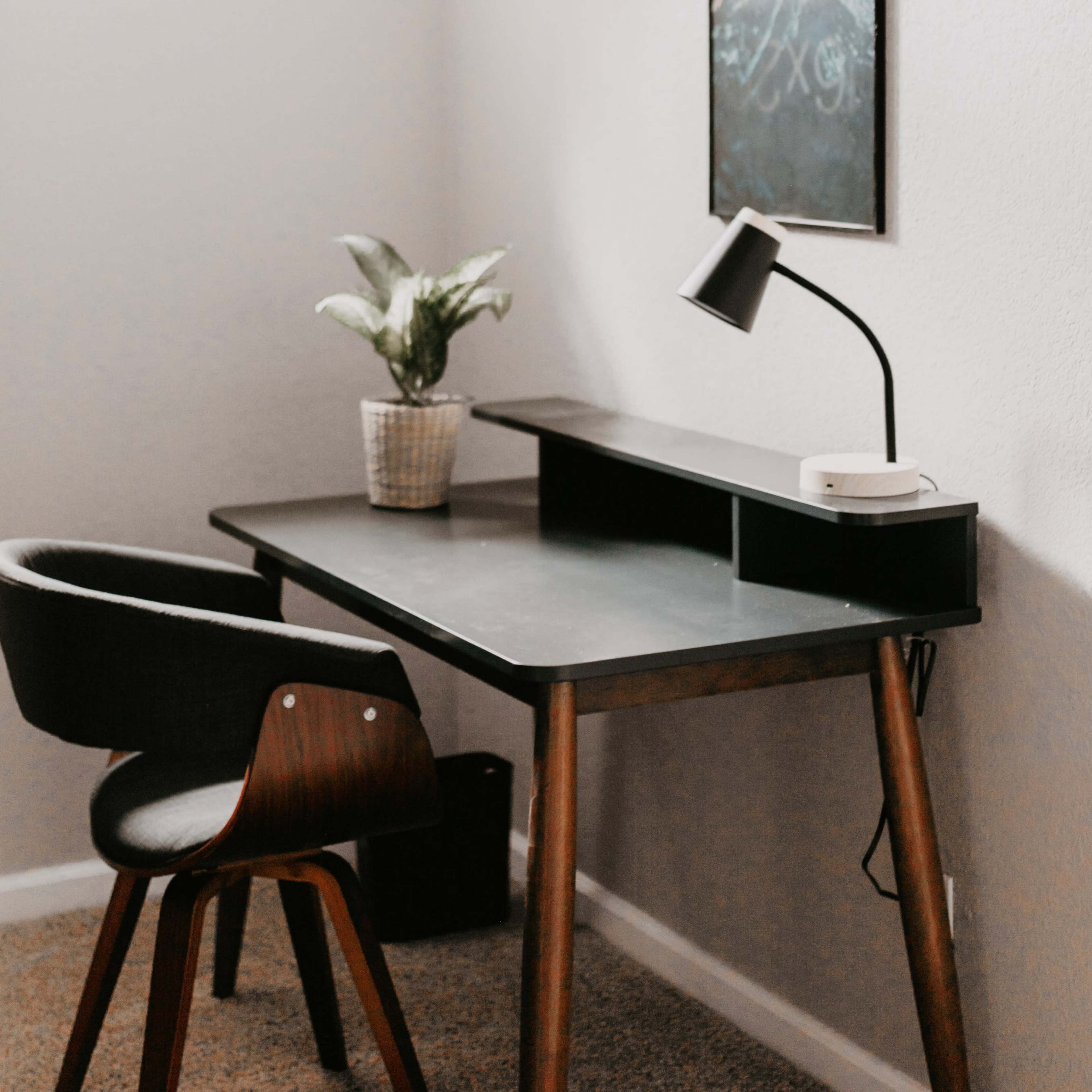 A modern desk with a chair, a small plant, and a lamp, positioned against a light-colored wall.
