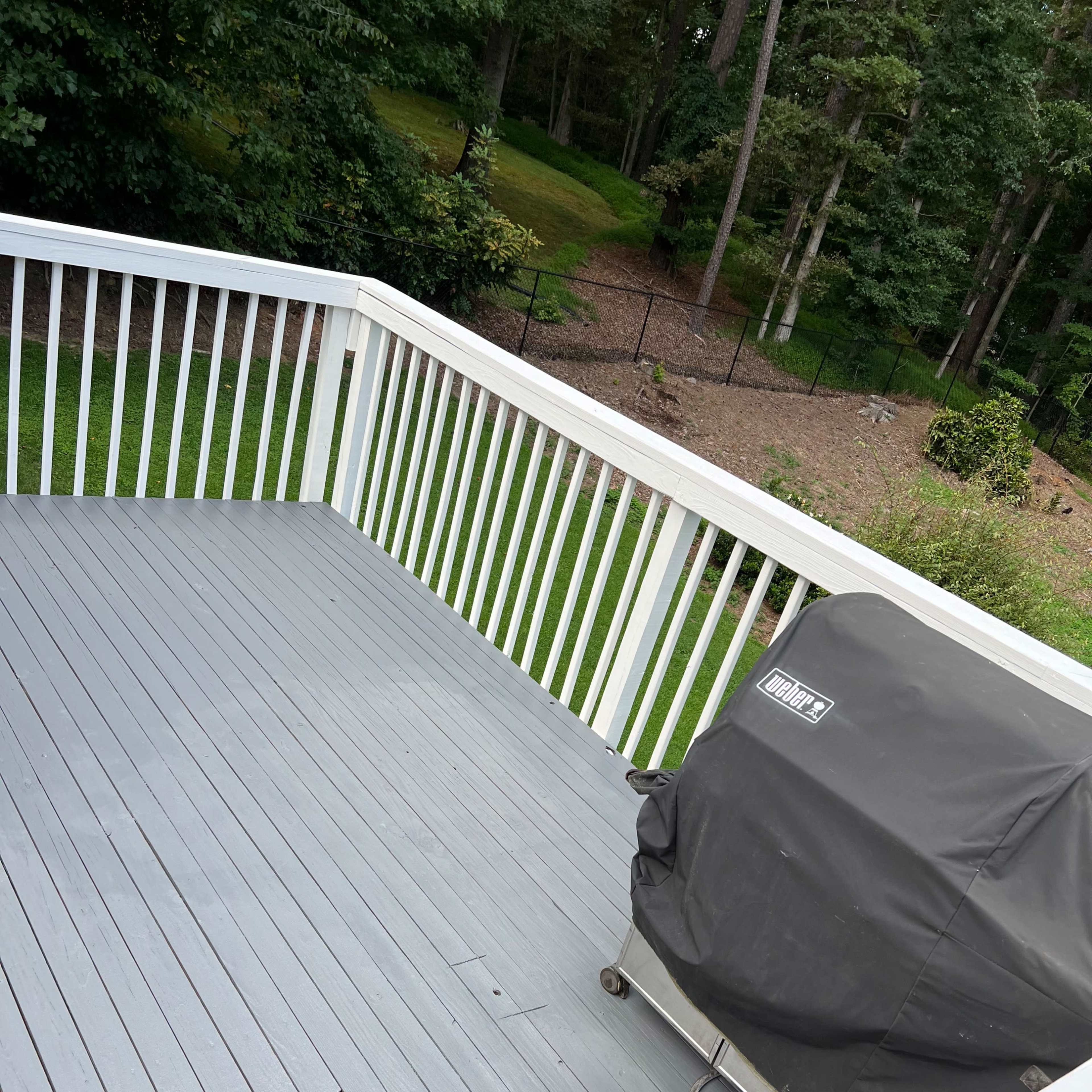 A gray wooden deck with a covered grill in one corner and a green lawn and trees visible beyond the railing.