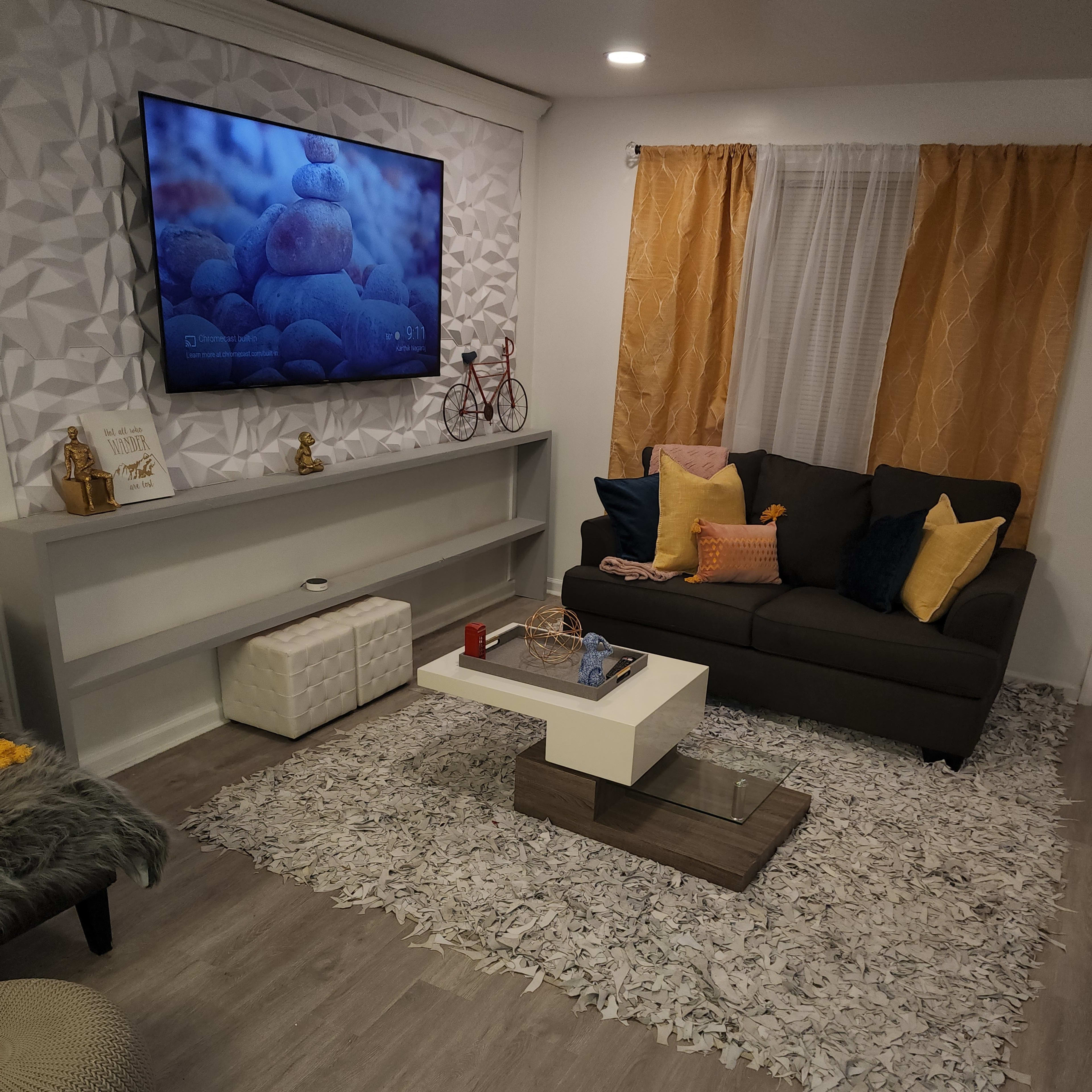 The room features a cozy seating area with a dark sofa, a coffee table, a TV mounted on the wall, and decorative elements like cushions and a rug.