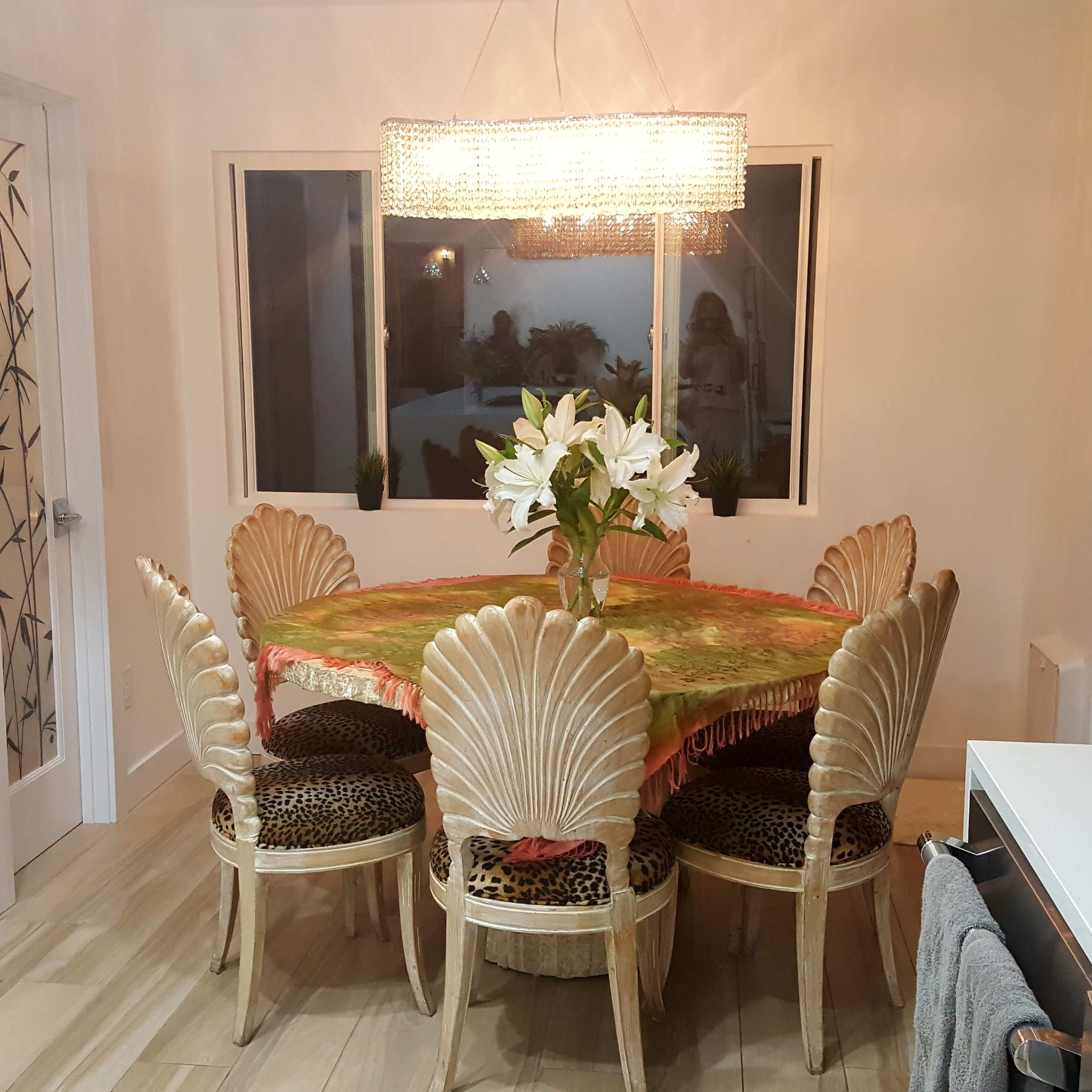 A round dining table with a colorful tablecloth is surrounded by ornate shell-patterned chairs in a well-lit room featuring large windows.