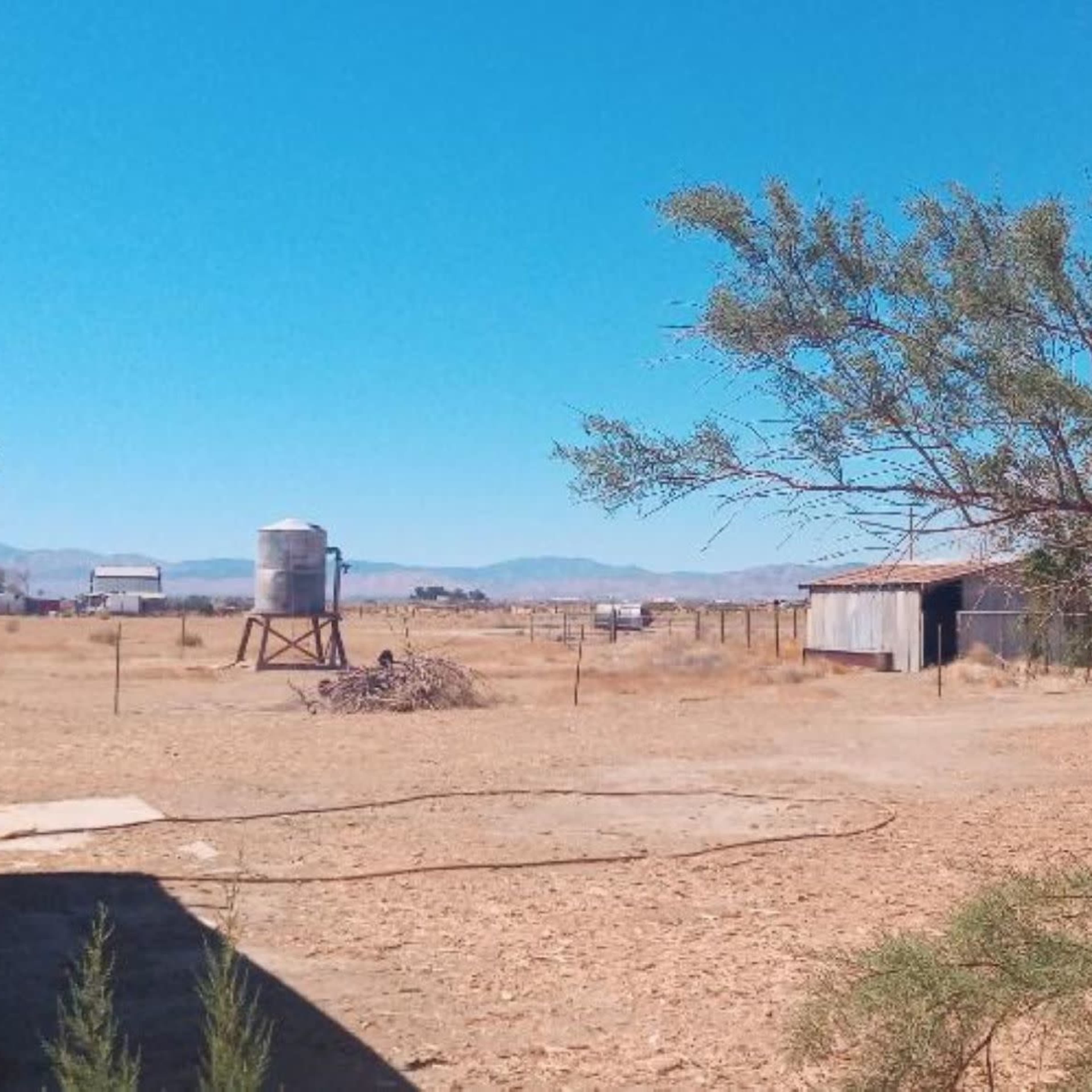 A rural scene featuring a dry landscape with a water tower, a barn, and sparse vegetation under a clear blue sky.