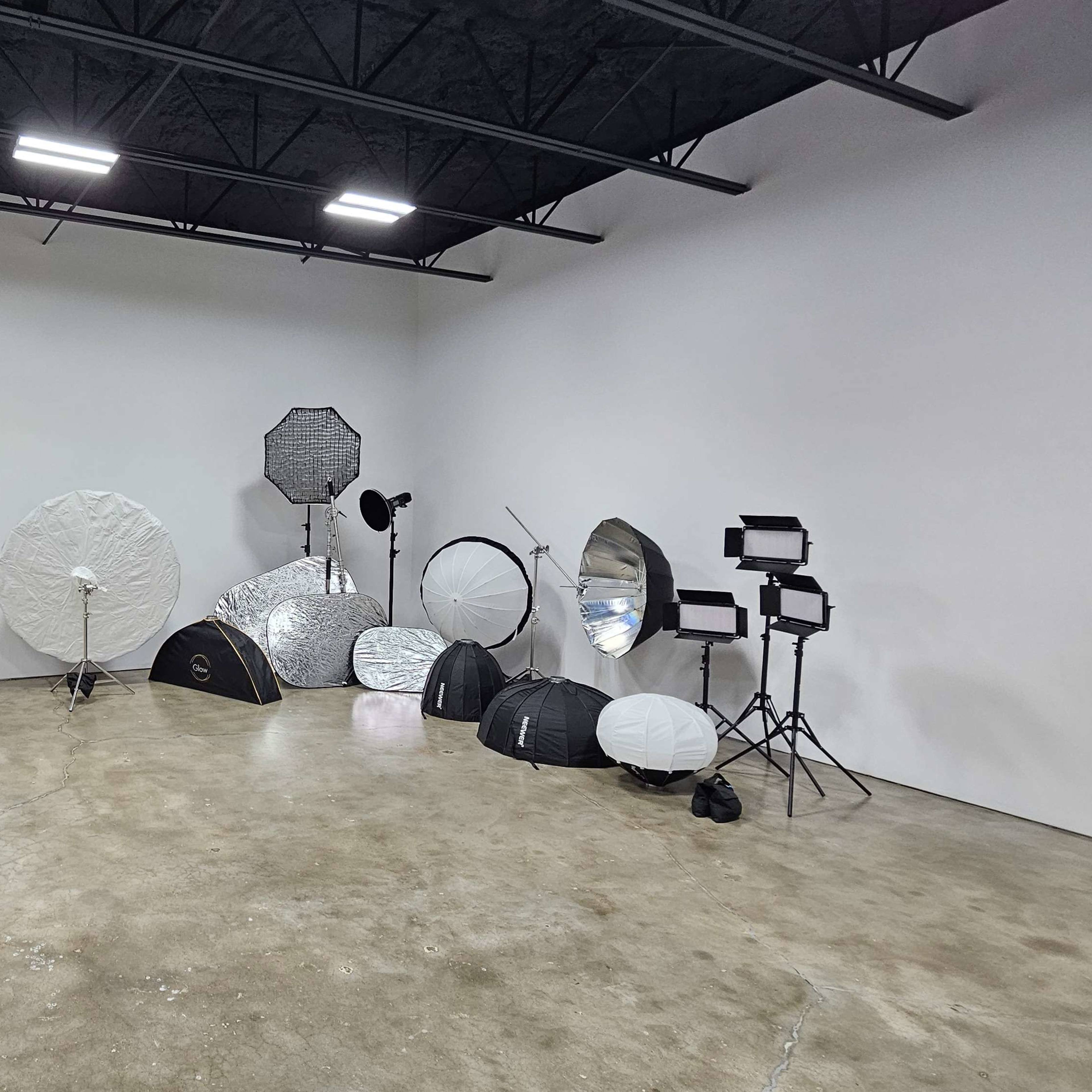 A spacious, empty studio featuring various photography equipment, including light reflectors, umbrellas, and softboxes arranged in the corner.