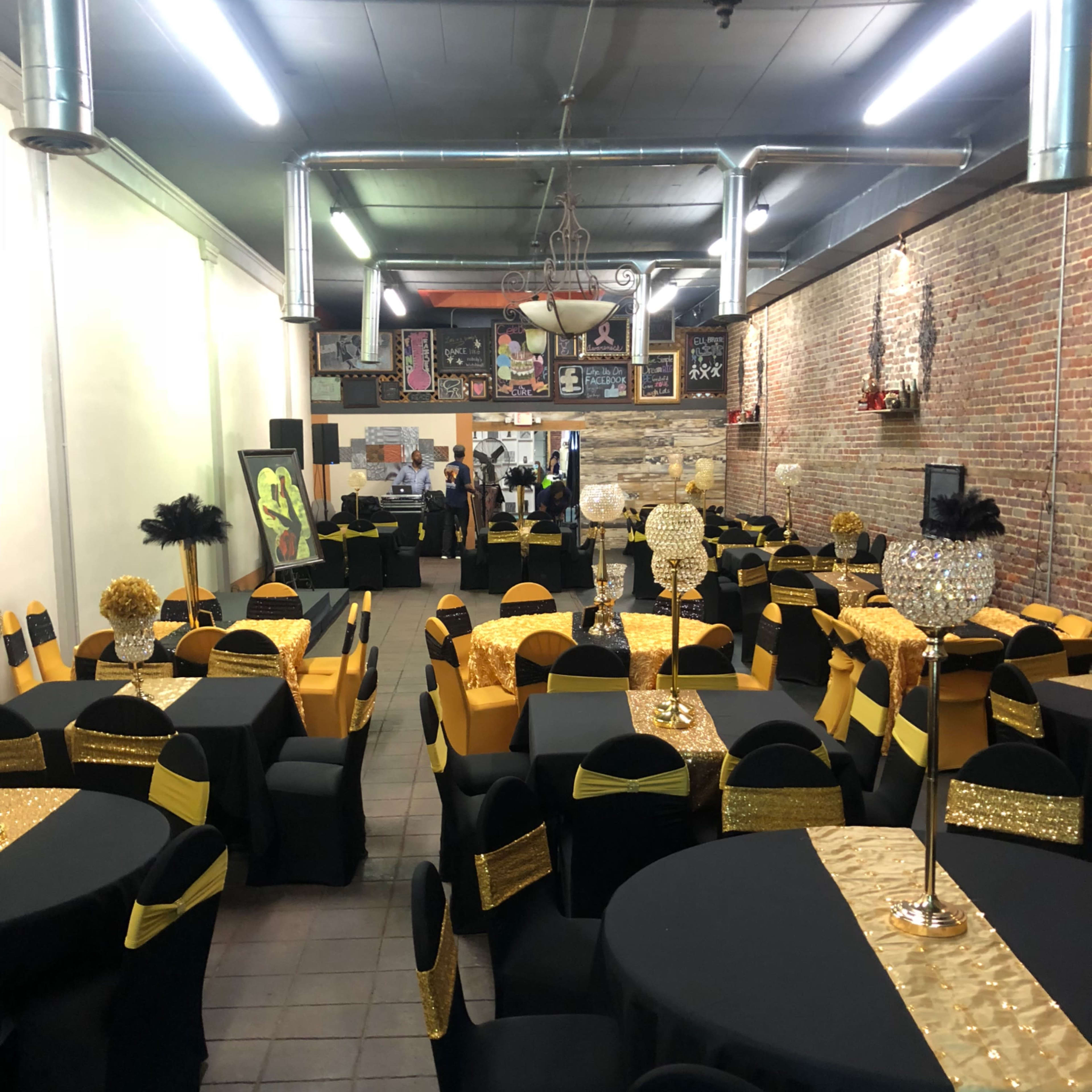 A large event space set up with black and gold tables and chairs, featuring decorative centerpieces and a brick wall in the background.