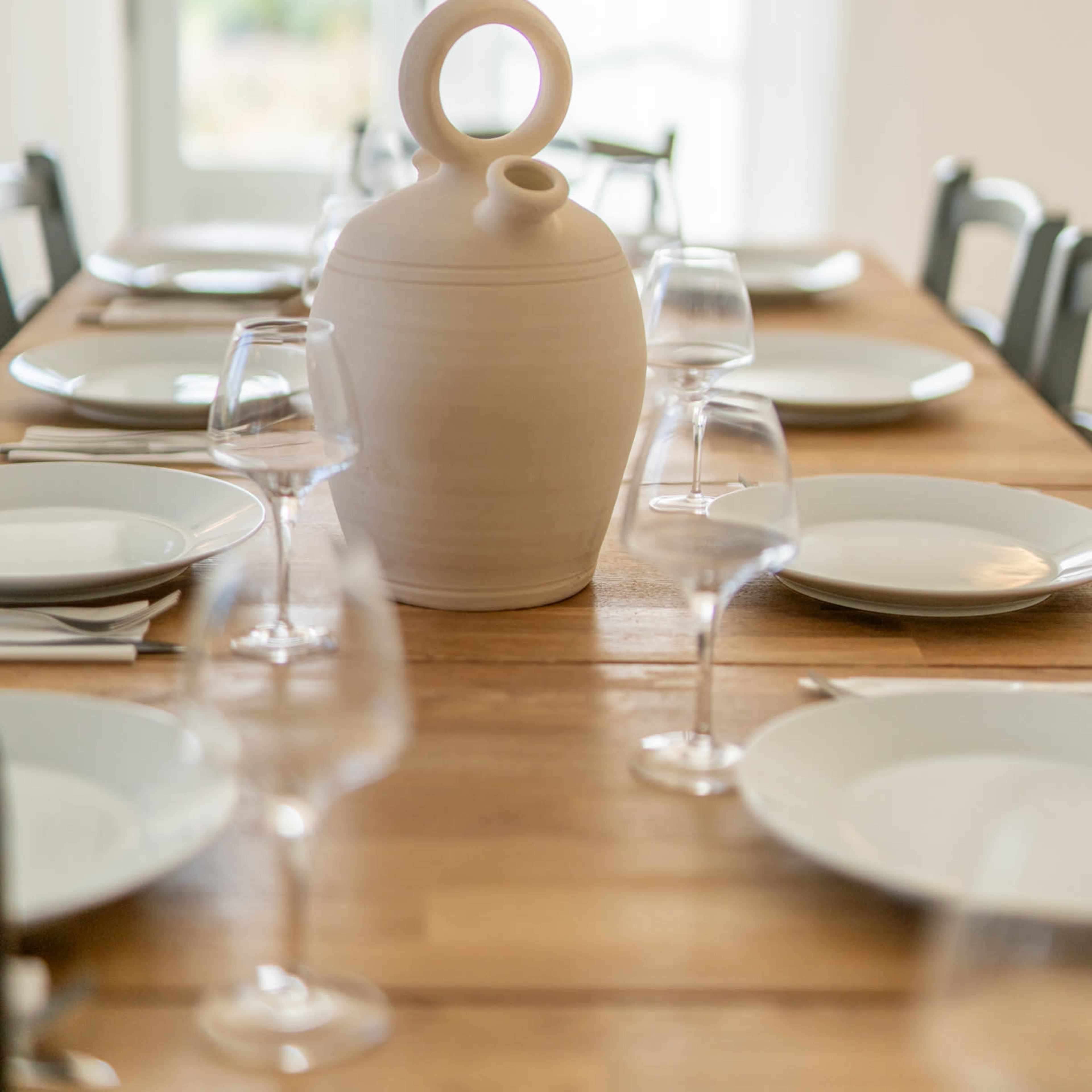 A large, light-colored ceramic jug sits on a wooden dining table set with white plates and wine glasses.