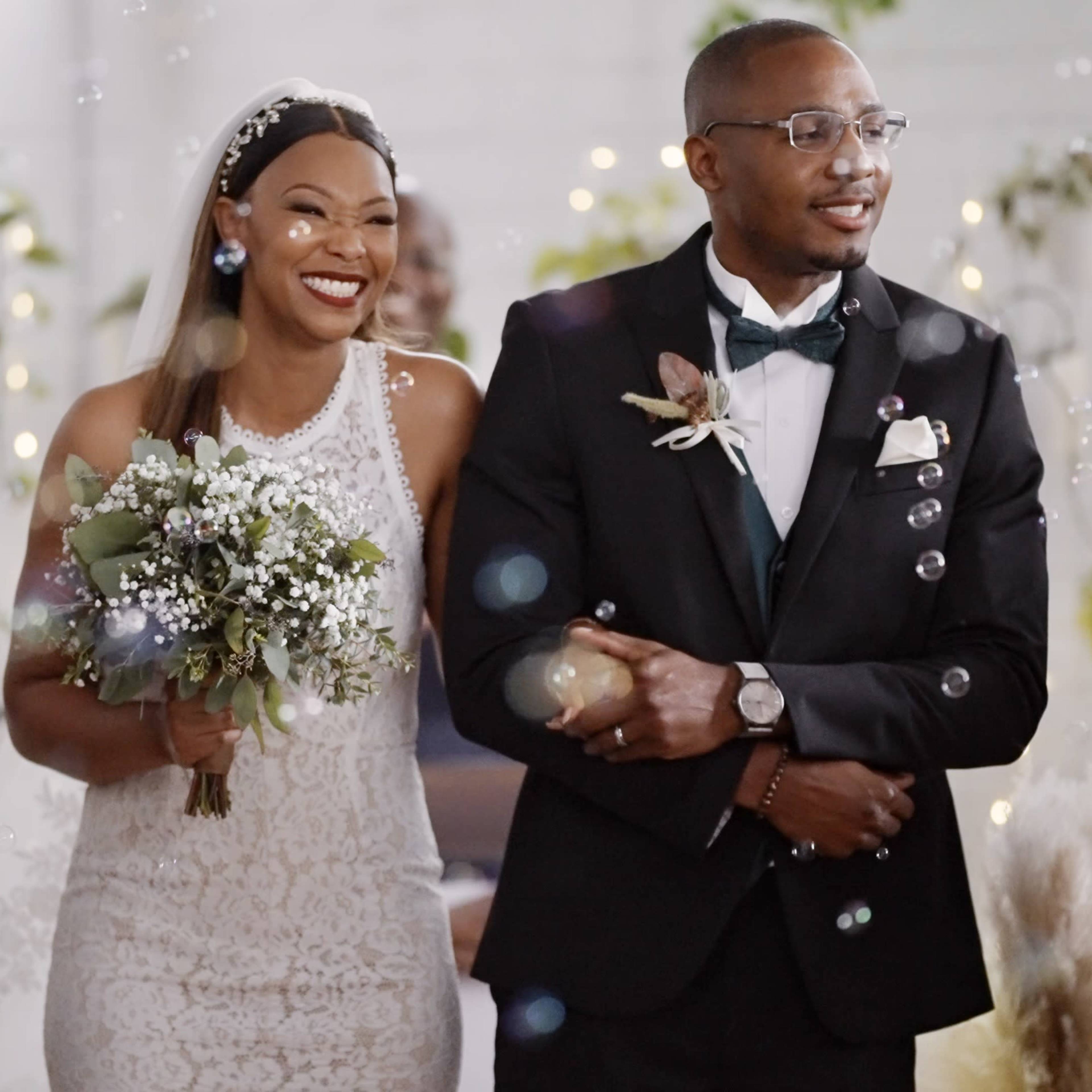 A bride in a lace wedding dress and a groom in a black tuxedo walk down the aisle, smiling and holding hands, with bubbles floating around them.