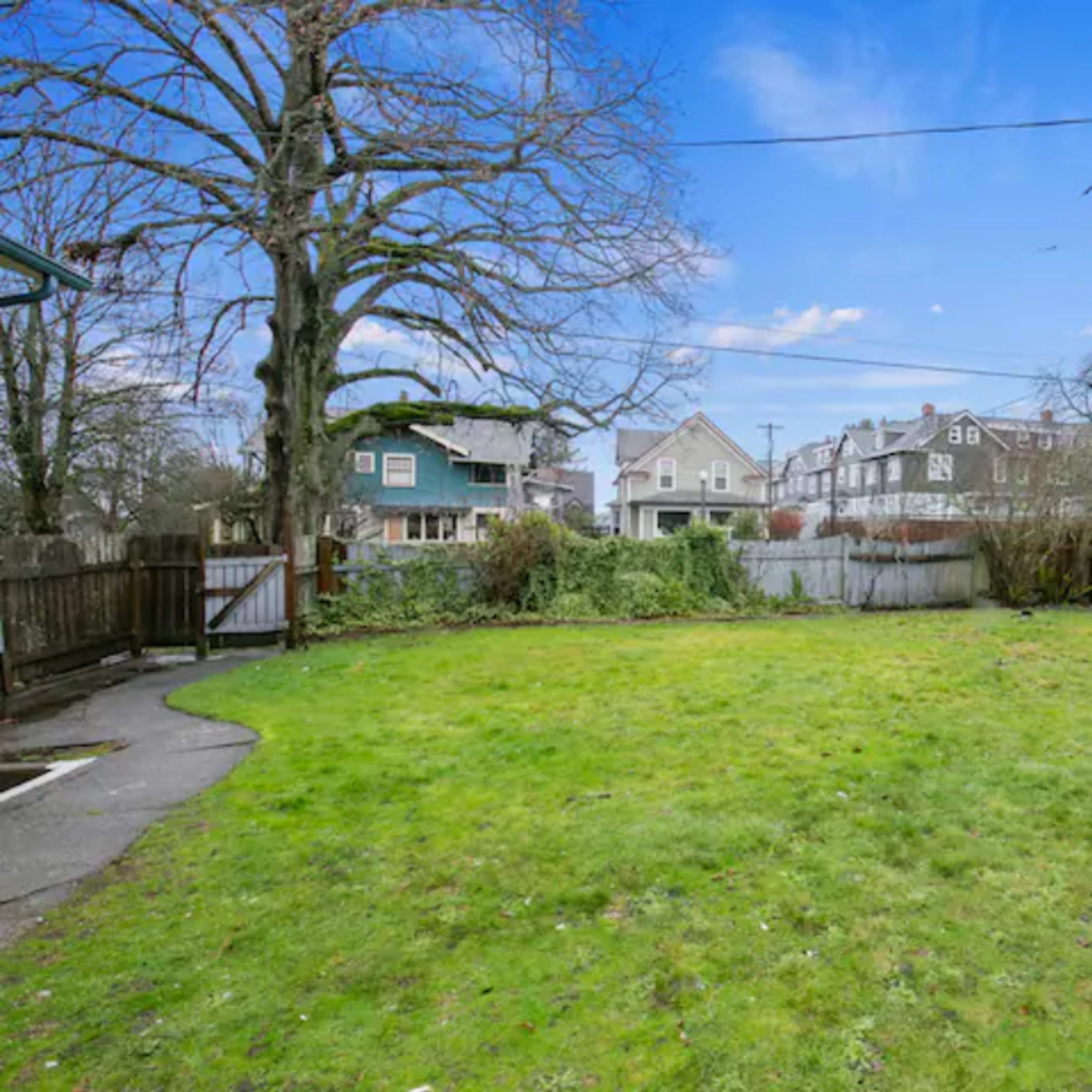 A spacious backyard with a grassy area, wooden fencing, and residential houses in the background under a clear blue sky.